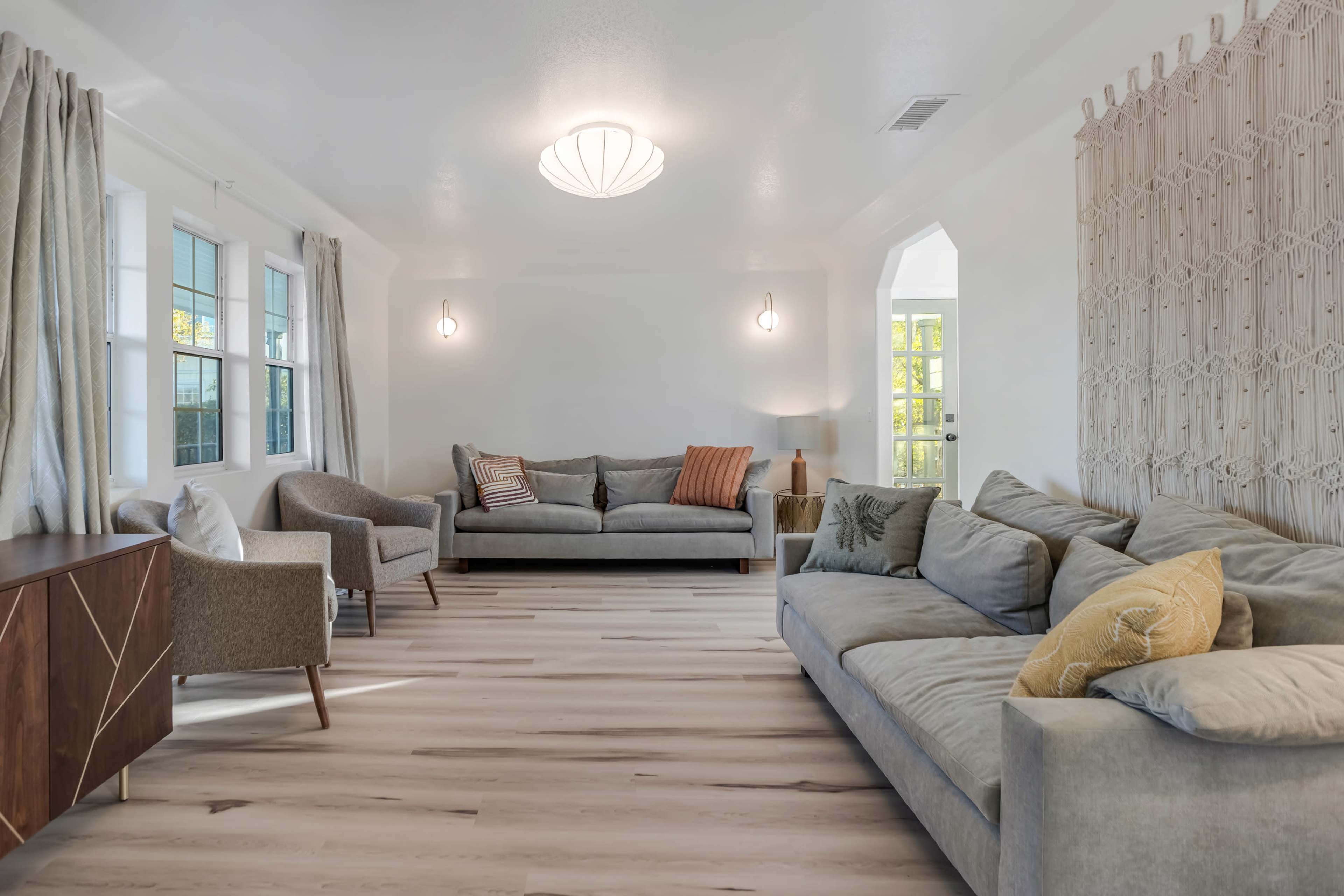 A bright living room with two gray sofas, a light-colored wooden floor, and large windows allowing natural light to enter.