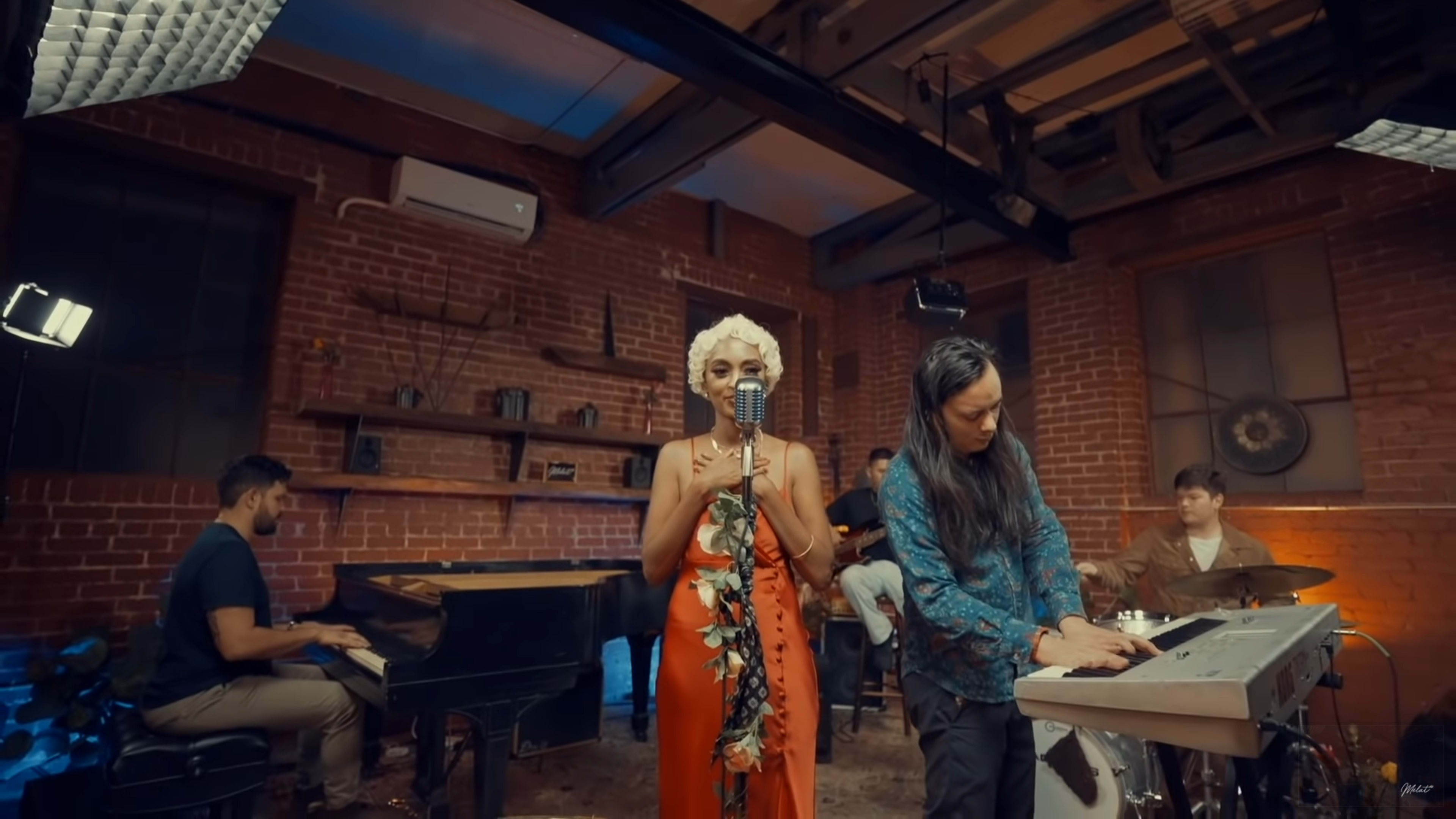 A vocalist stands in front of a vintage microphone while a musician plays a keyboard, and additional band members are positioned at a piano and drums in a rustic studio setting with exposed brick walls.