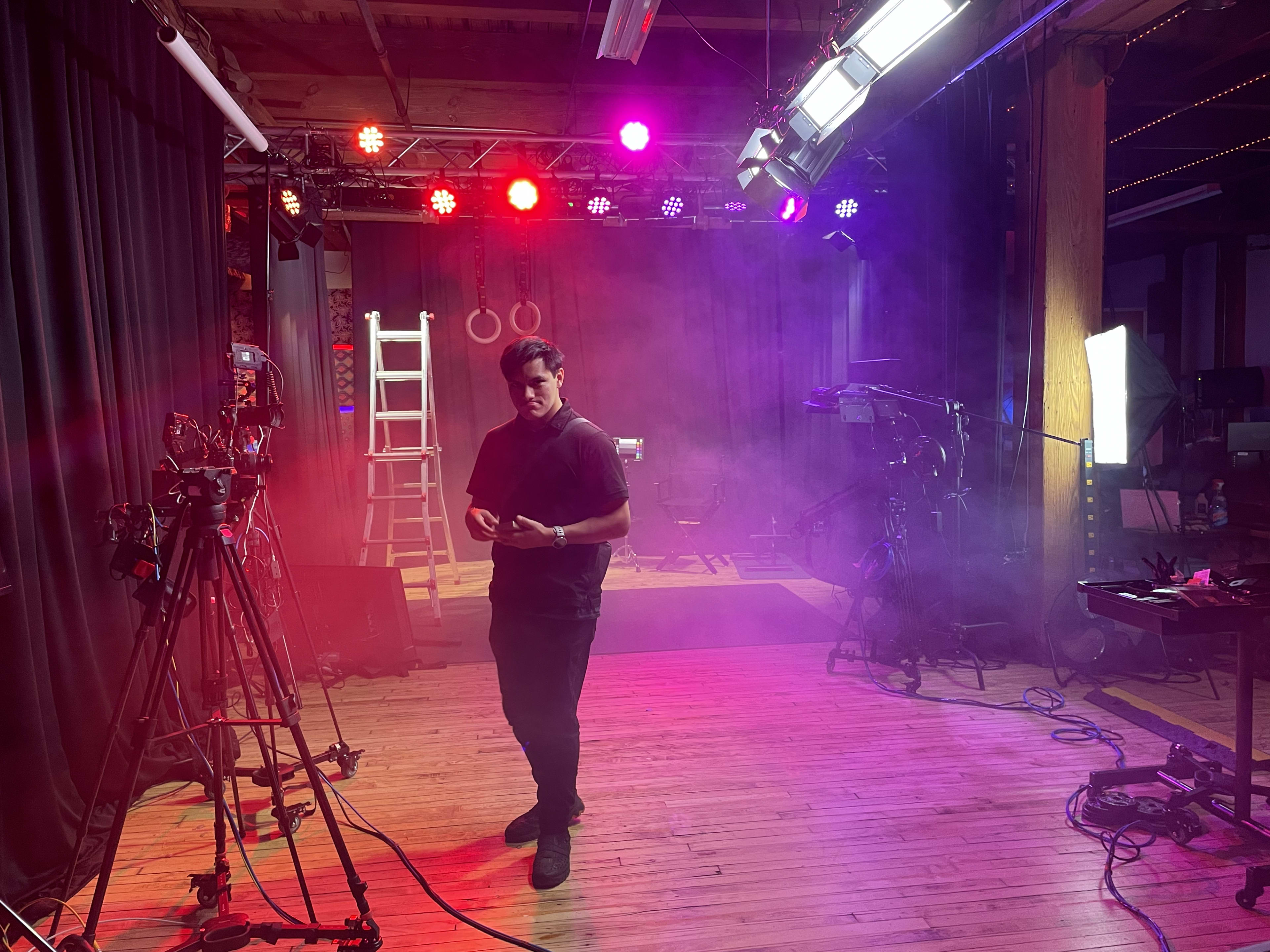 A man stands in a dimly lit studio filled with colorful stage lights, with cameras and a ladder in the background.