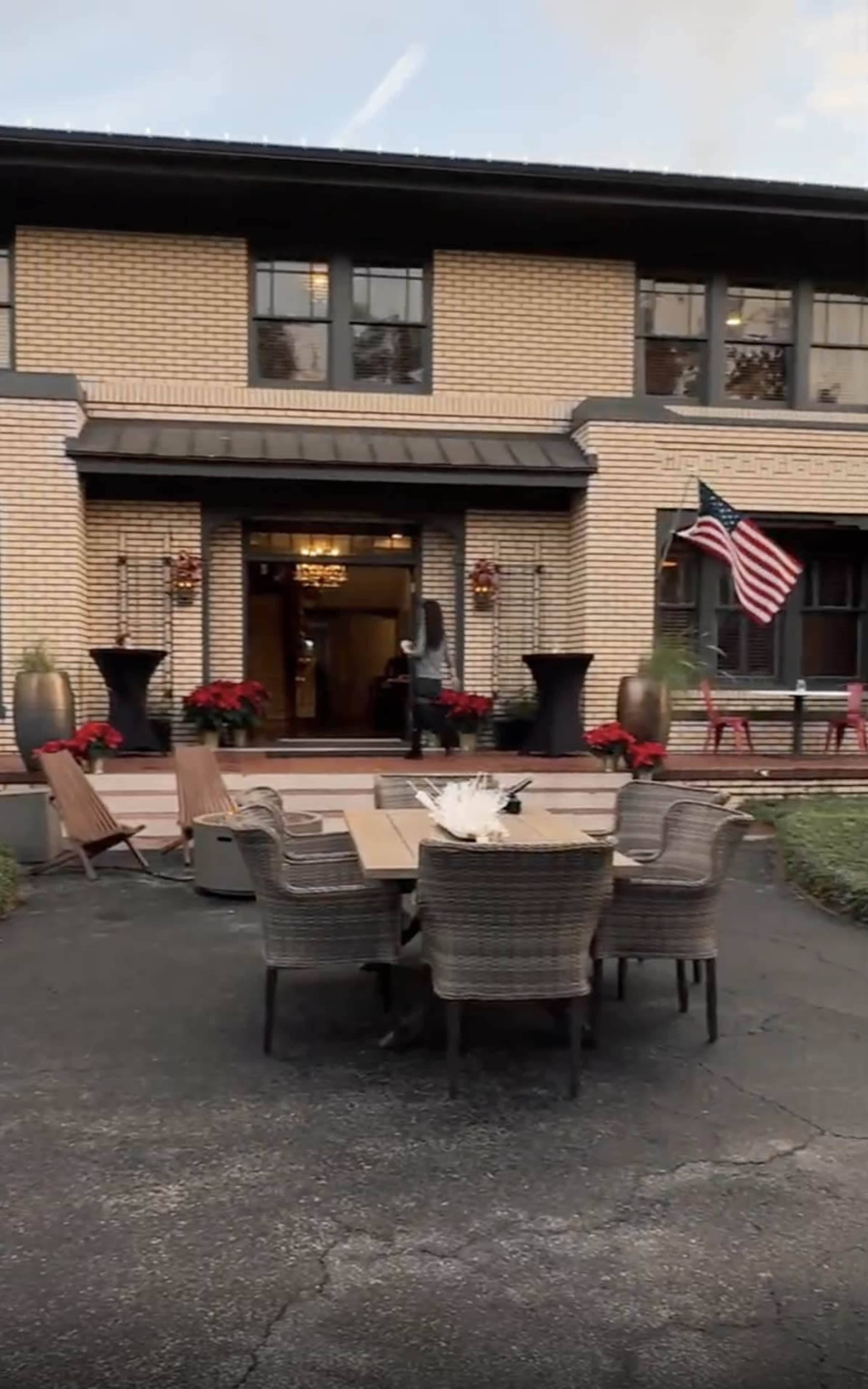A spacious outdoor dining area with a large table and wicker chairs is set in front of a brick building, adorned with holiday decorations and an American flag.