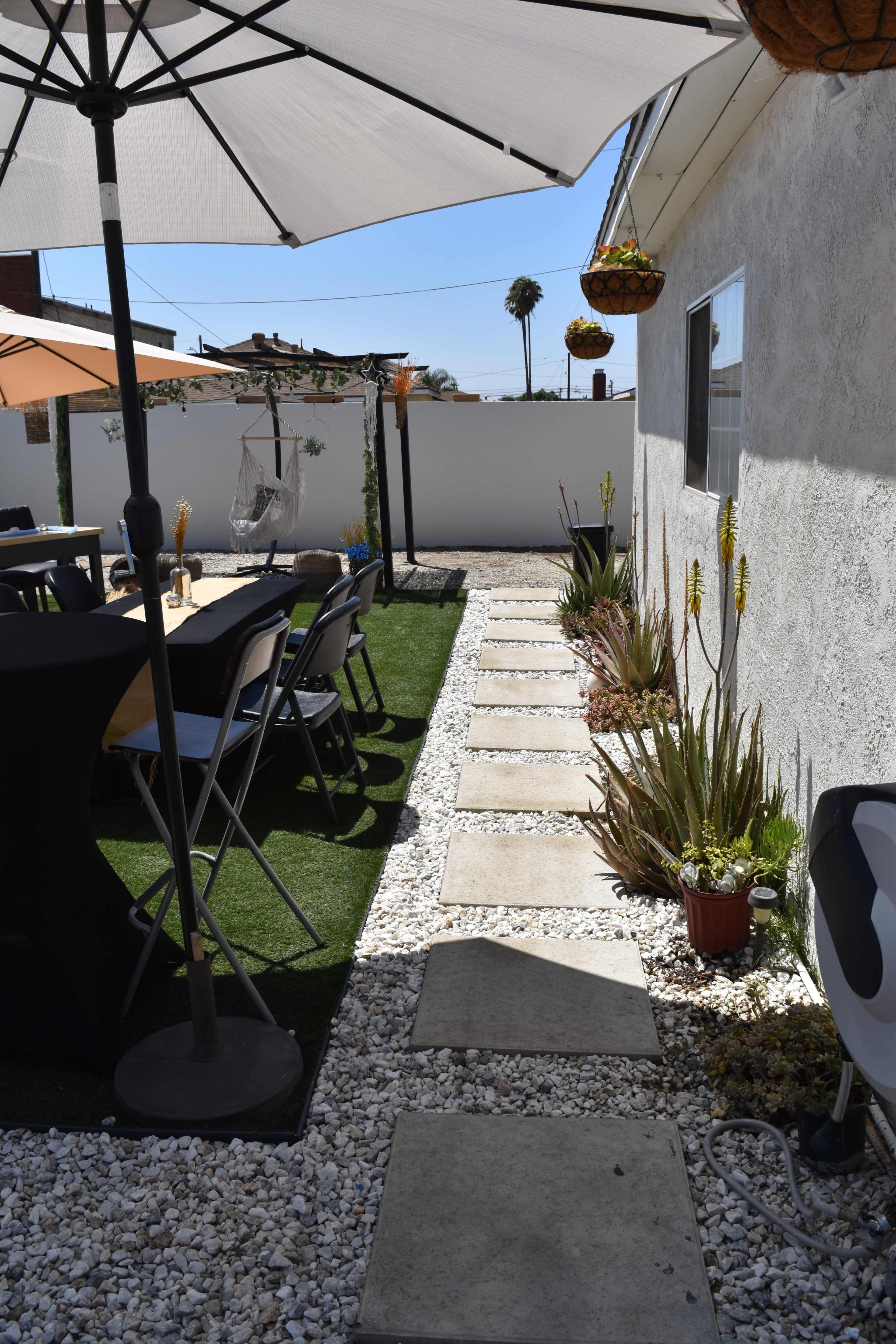 A pathway of stone slabs leads through a neatly arranged garden with gravel and potted plants beside a white wall.