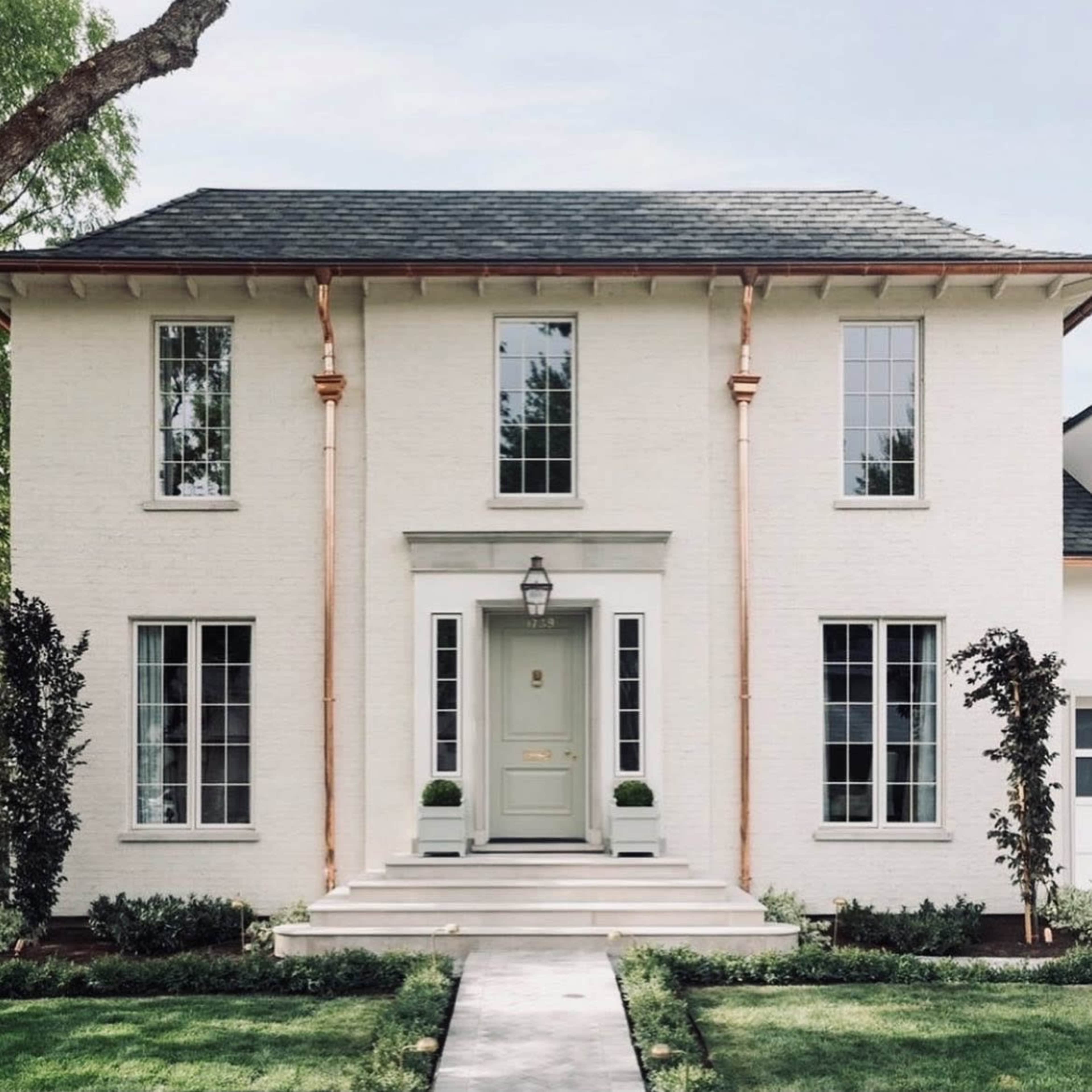 A two-story white brick house features a central door with a light-colored frame, flanked by large windows and surrounded by neatly trimmed landscaping.