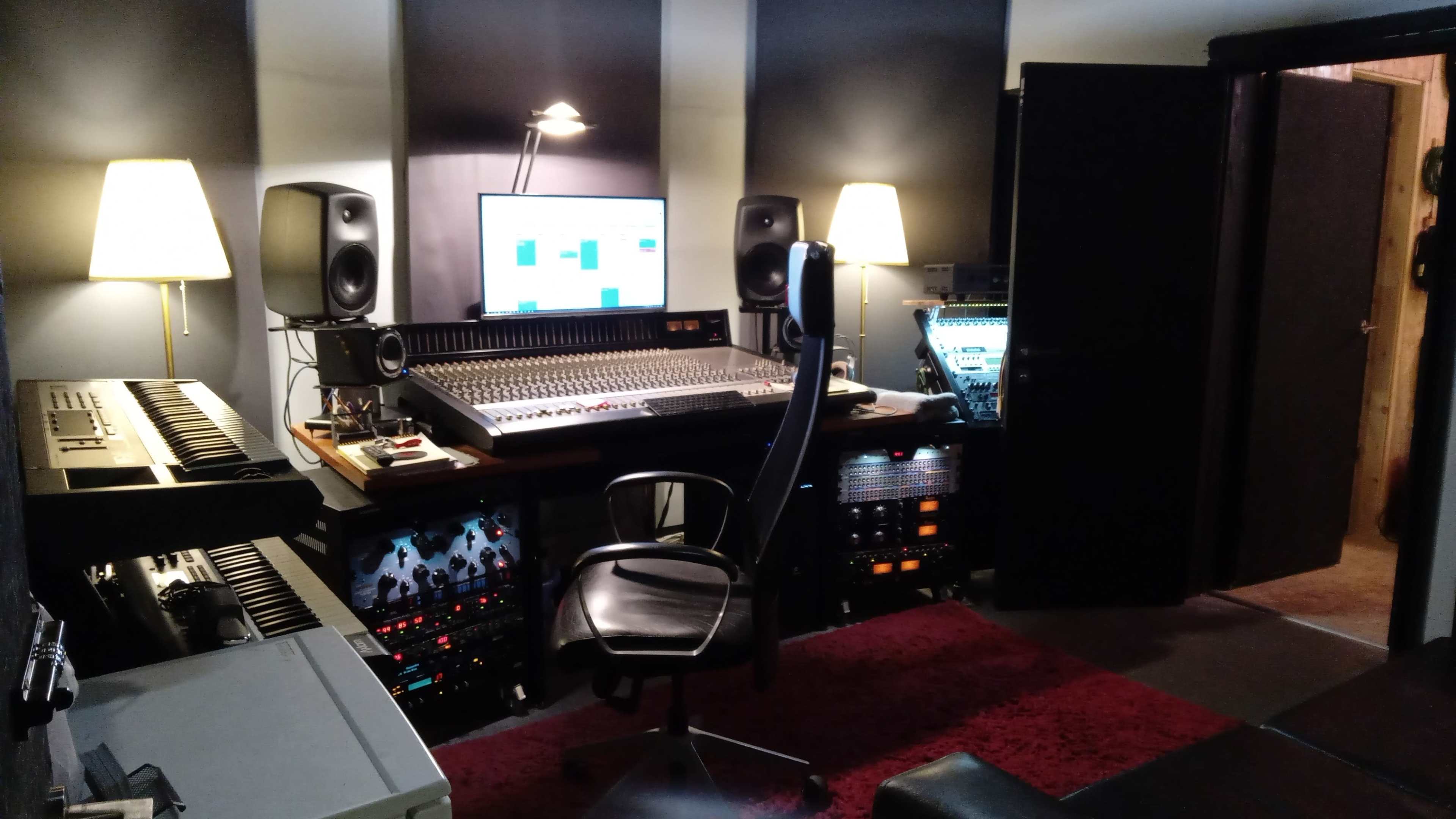 A music studio featuring a mixing console, monitors, a keyboard, and various audio equipment in a dimly lit room with a chair and carpets.