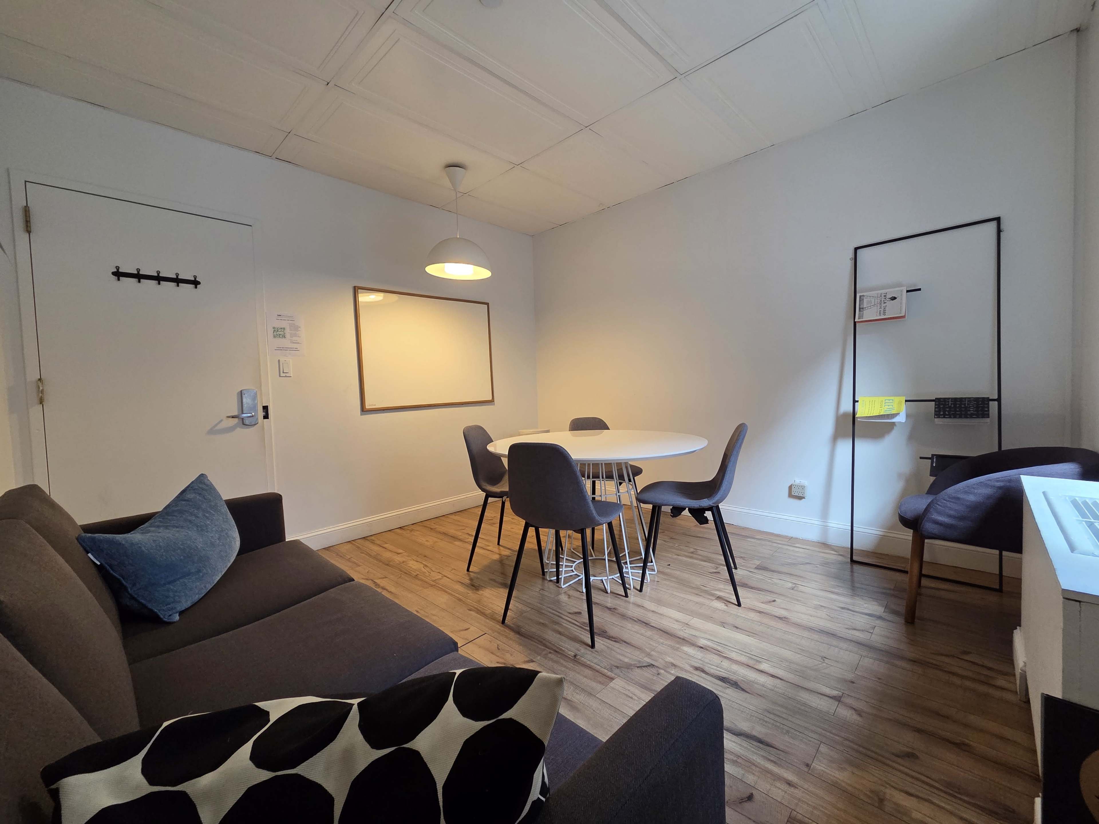 The image shows a small, modern room with a circular dining table surrounded by six chairs, a sofa with a patterned cushion, and a whiteboard on the wall.