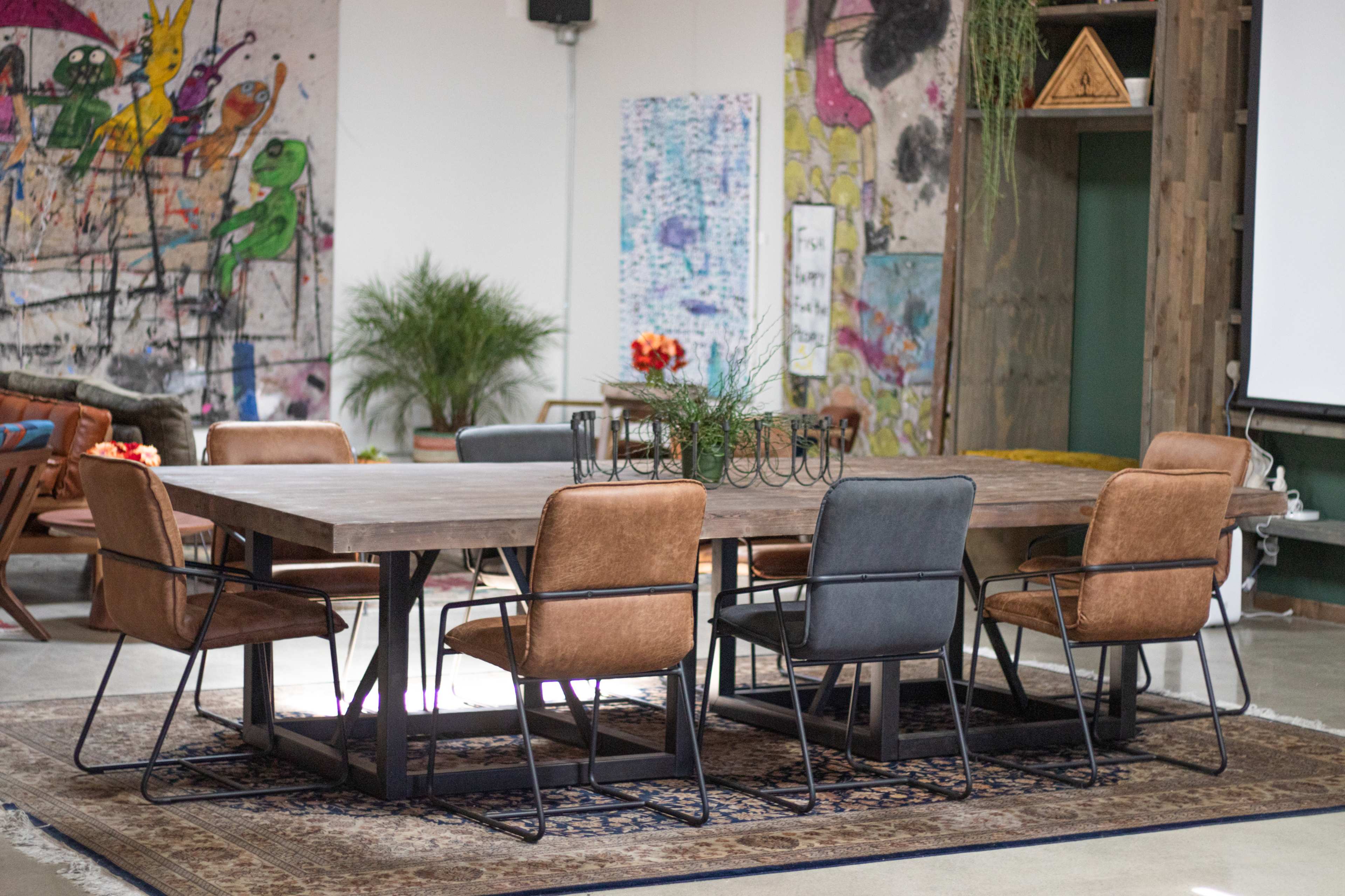 The image shows a large wooden dining table surrounded by six chairs on a decorative rug, set against a colorful graffiti-style wall.