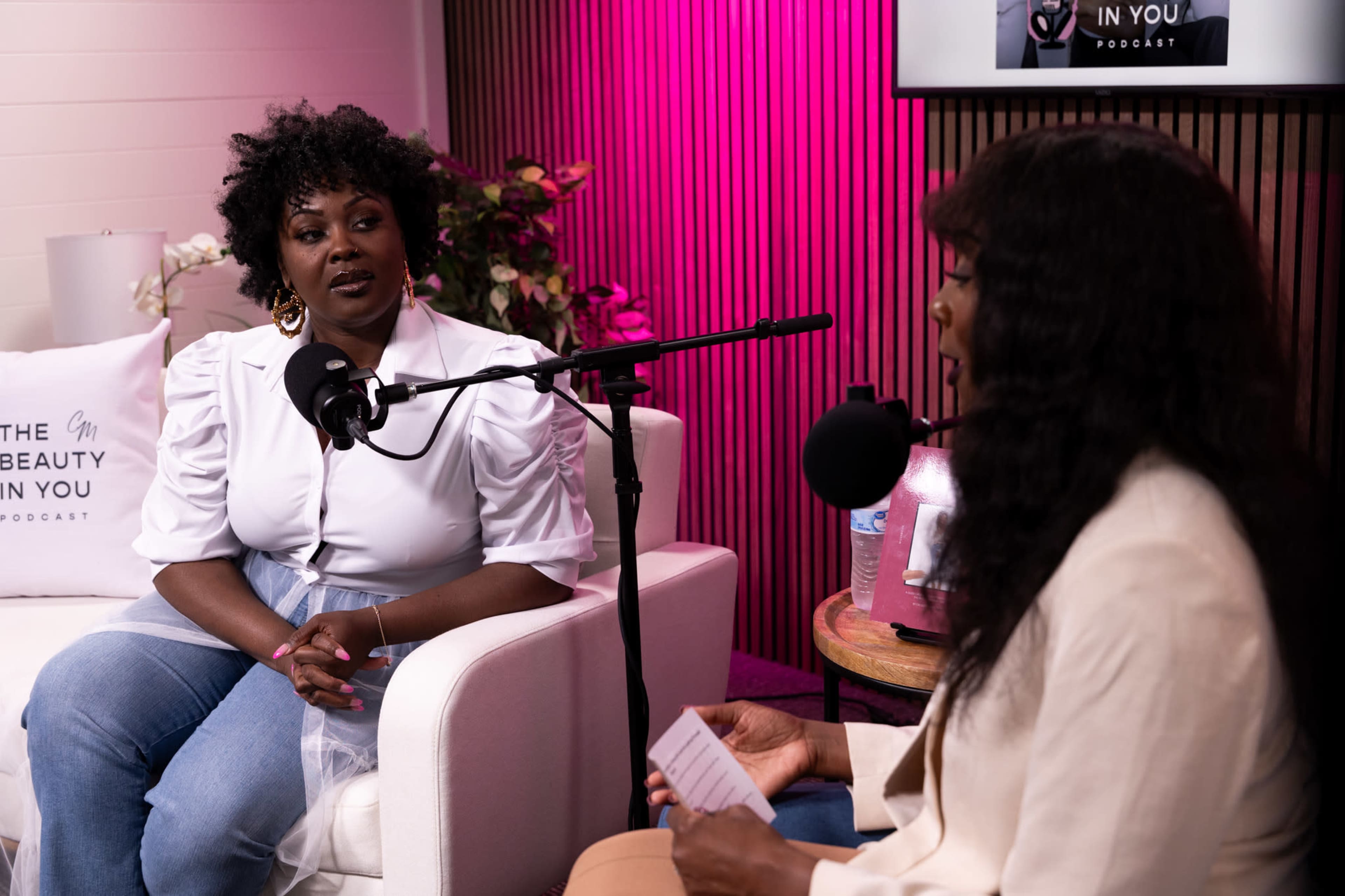 Two women sit in a podcast studio, engaging in conversation, with one woman on a white couch and the other speaking into a microphone.