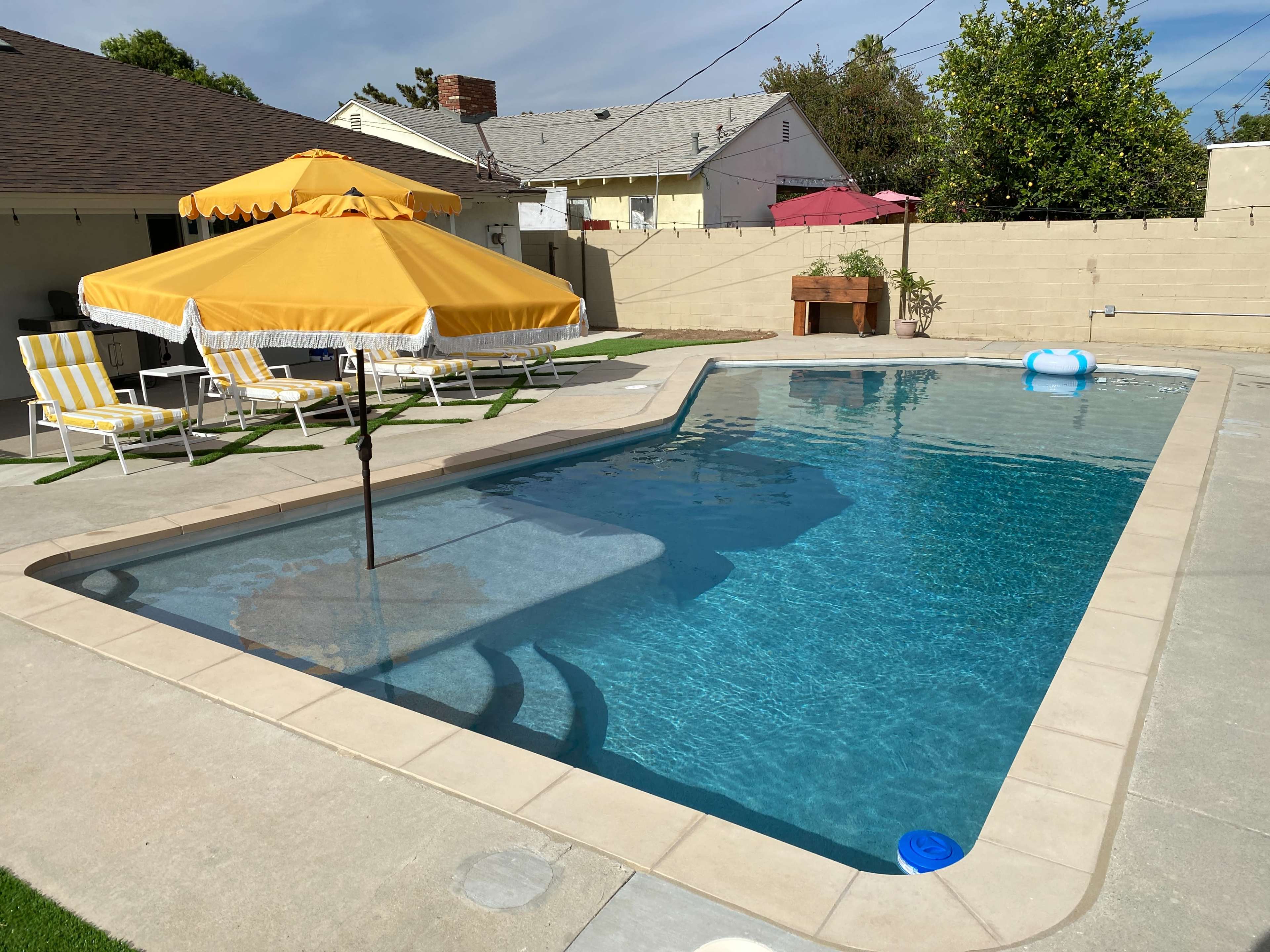 A rectangular swimming pool surrounded by lounge chairs and a yellow umbrella in a residential backyard.