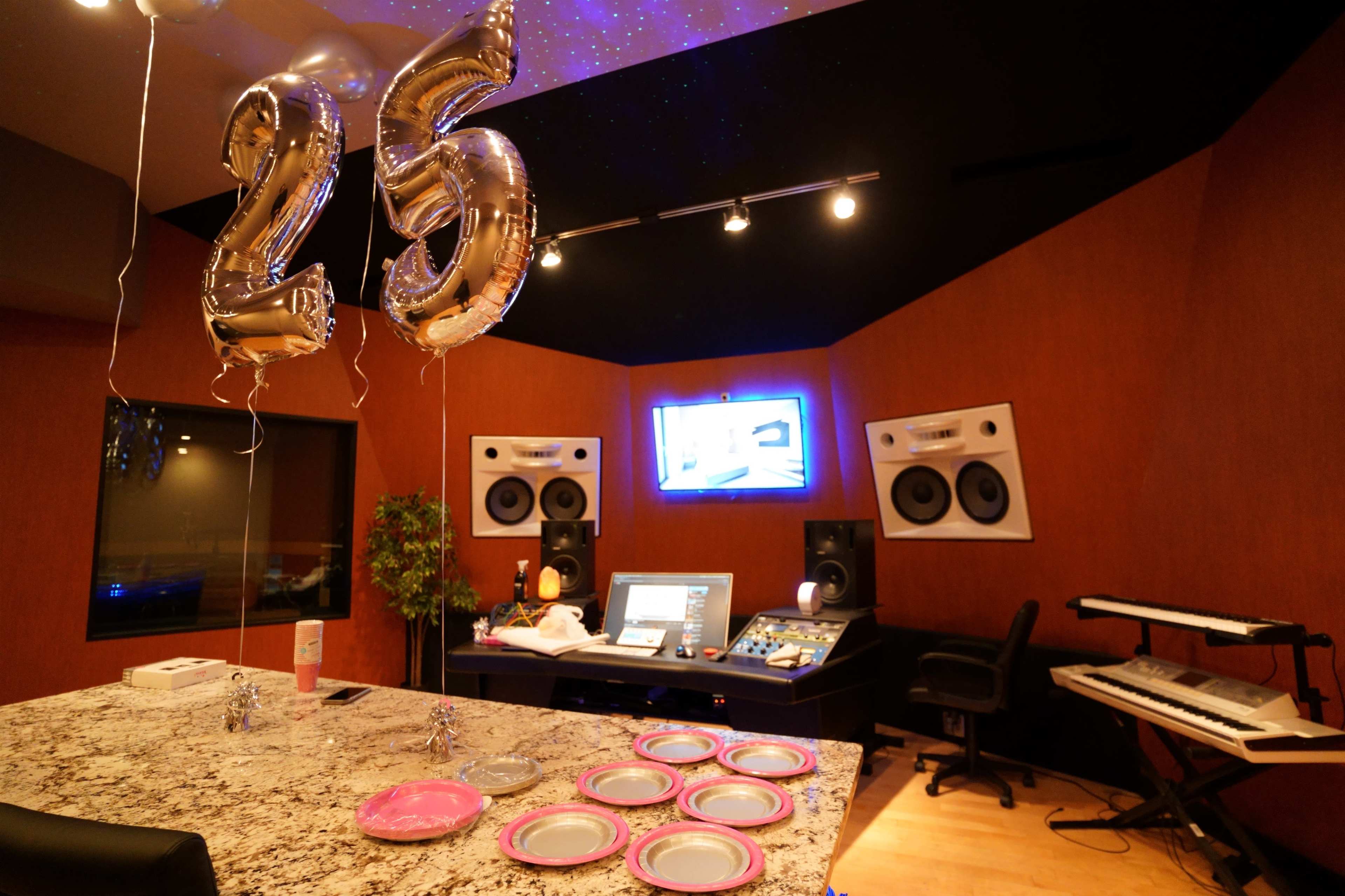 A music studio with "25" balloons, a decorated table with pink plates, and audio equipment in the background.