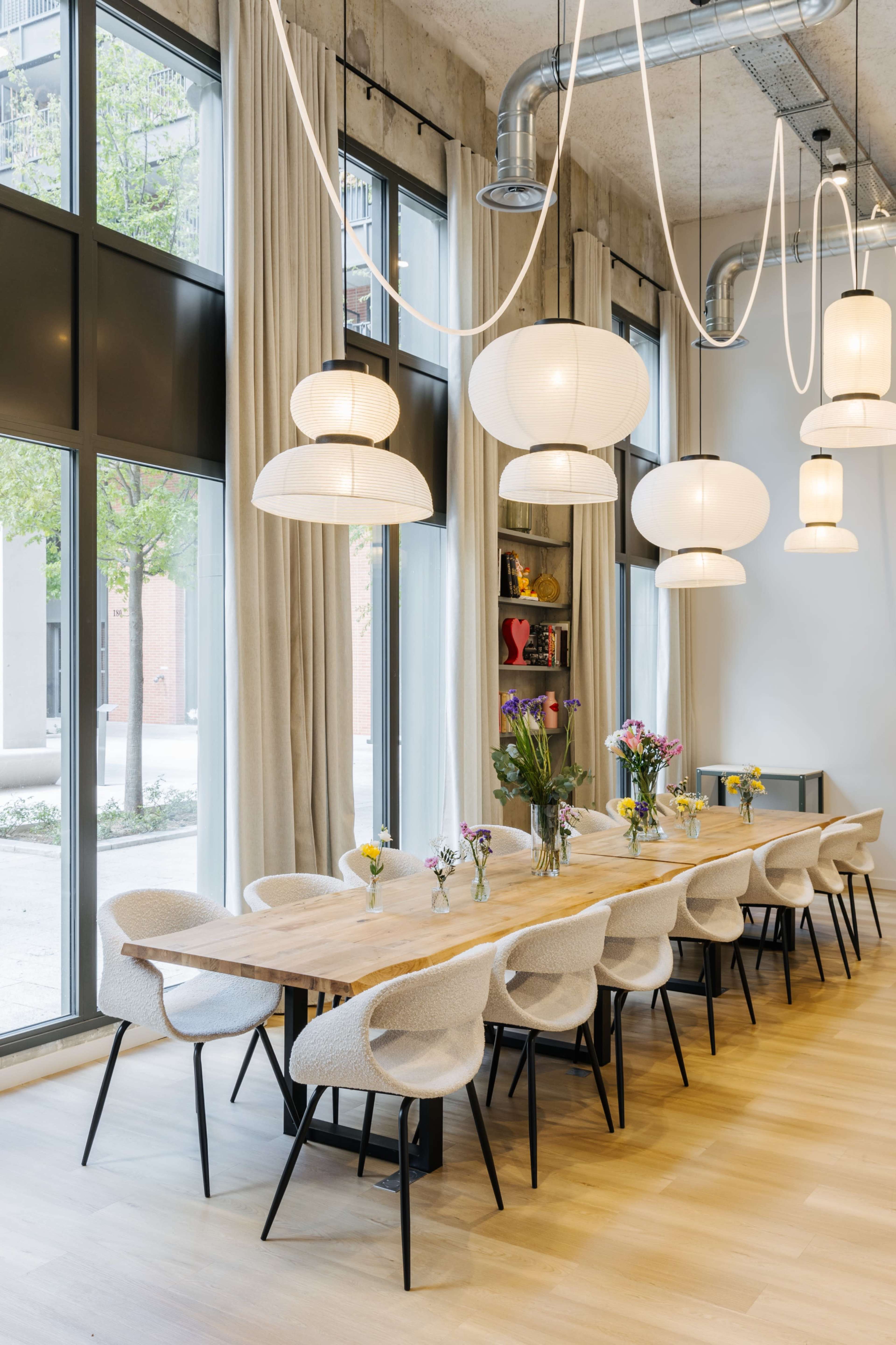 A long wooden dining table is surrounded by upholstered chairs in a bright room illuminated by large pendant lights and natural light from floor-to-ceiling windows.