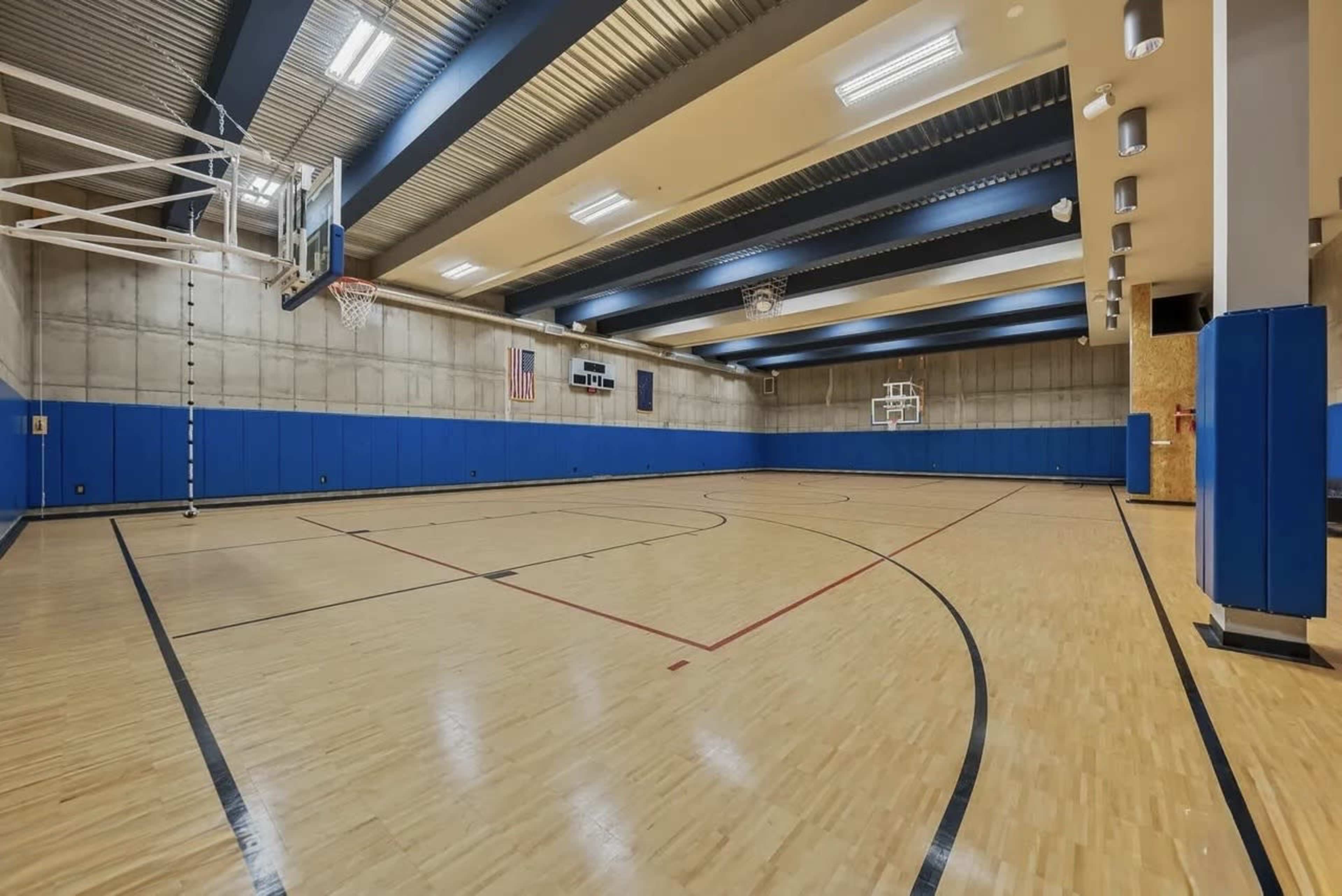 The image shows an empty indoor basketball court with wooden flooring, basketball hoops at each end, and blue wall padding.