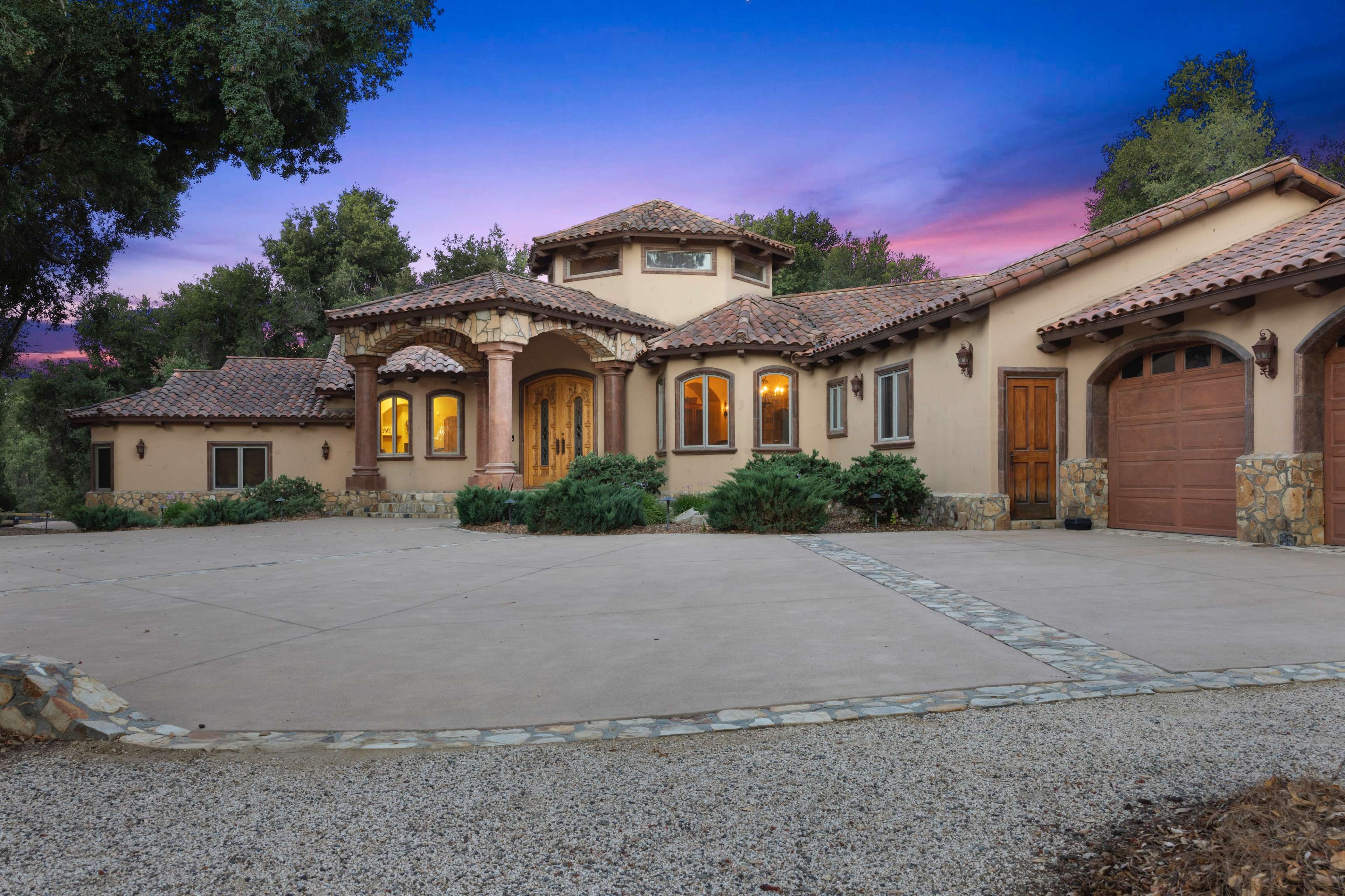 The image shows a large, custom-designed house with a tile roof, stone accents, and a paved driveway surrounded by greenery, set against a colorful sunset backdrop.