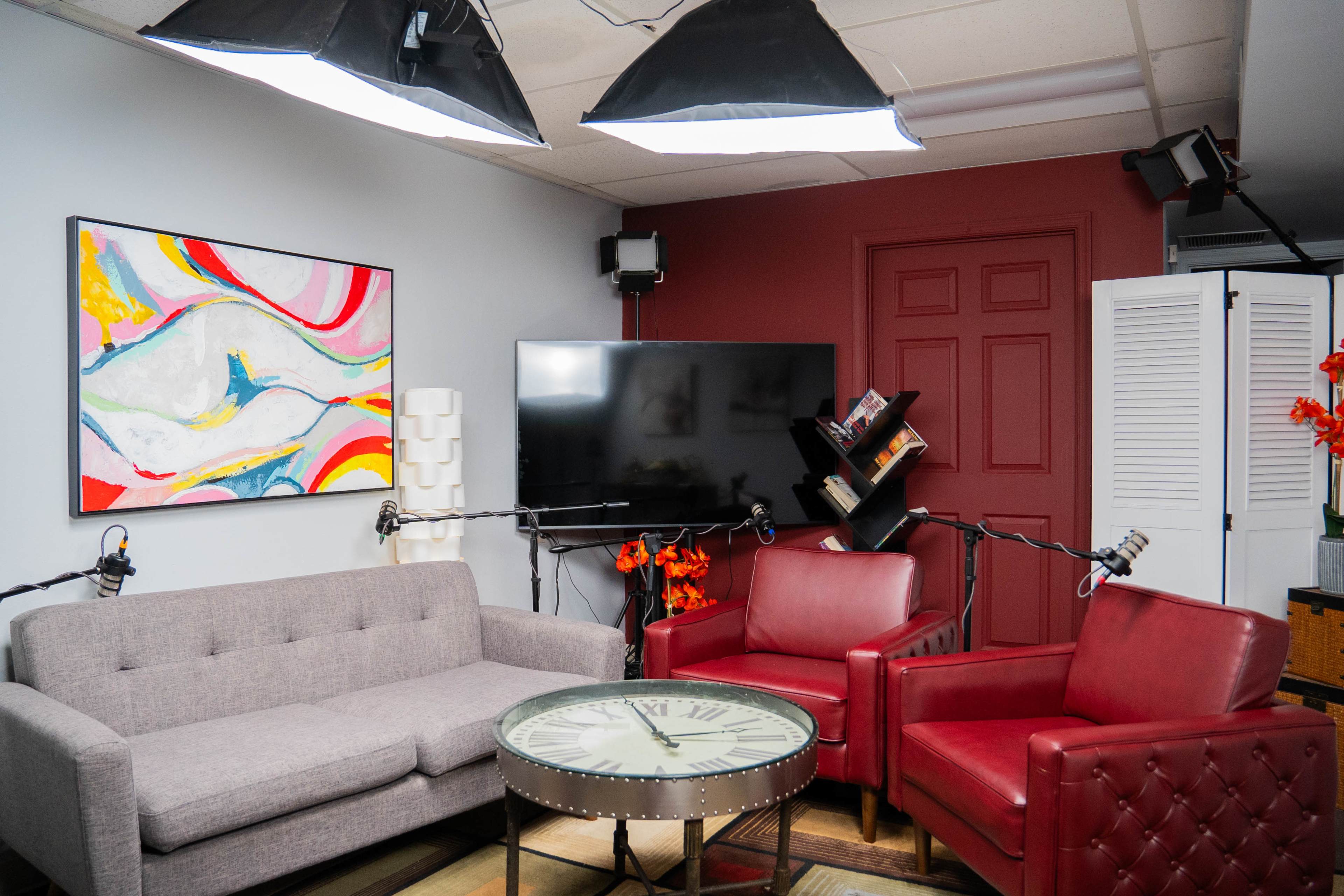 A modern studio space featuring a gray sofa, two red chairs, a round table with a clock design, and a large television on the wall, all illuminated by overhead lights.