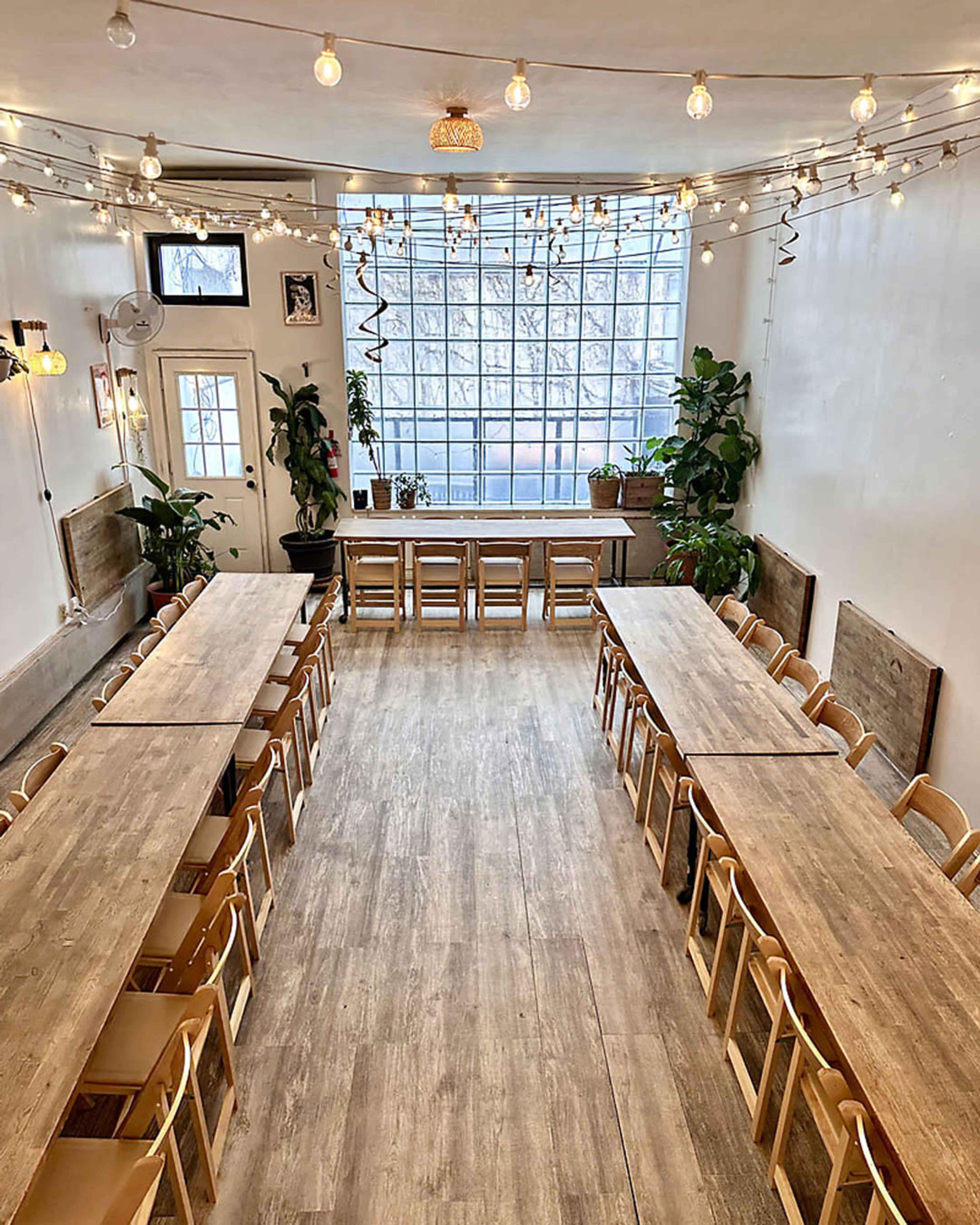 The image shows a spacious room with wooden tables arranged in a U-shape, surrounded by chairs, and illuminated by string lights.