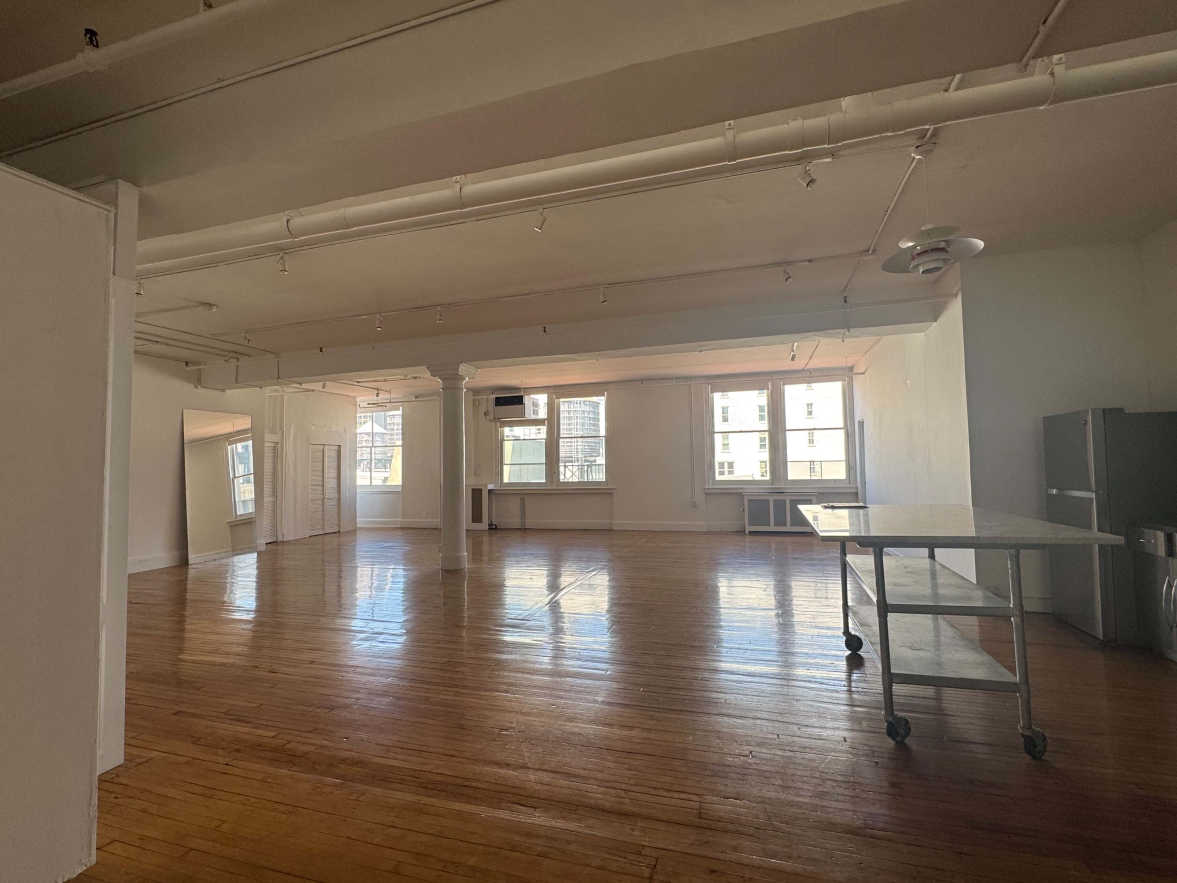 The image shows a spacious, empty room with wooden flooring, large windows, and minimalist furnishings.