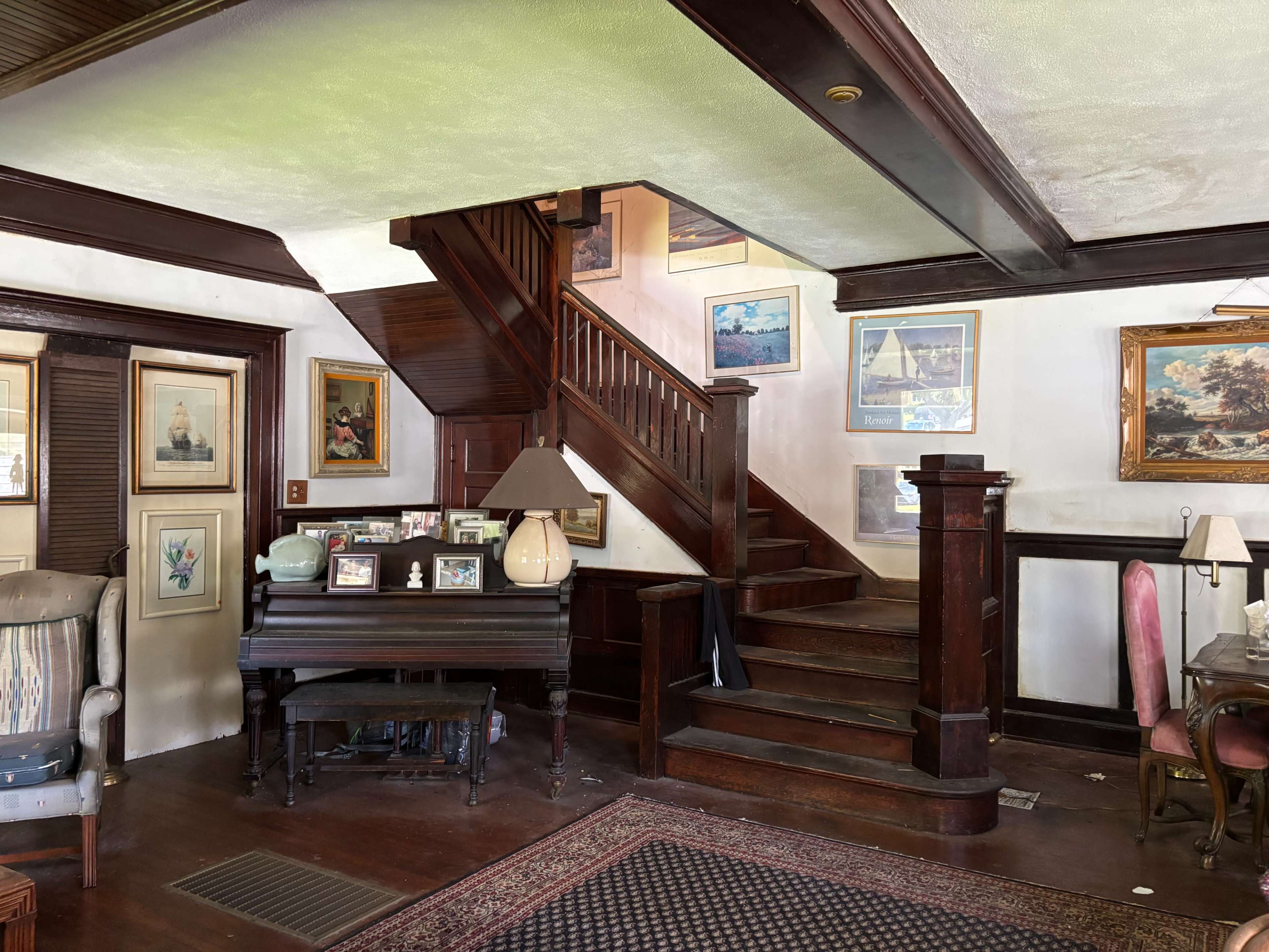 The image shows a cozy living space with a staircase, framed artworks on the walls, and a piano beside a collection of photographs.