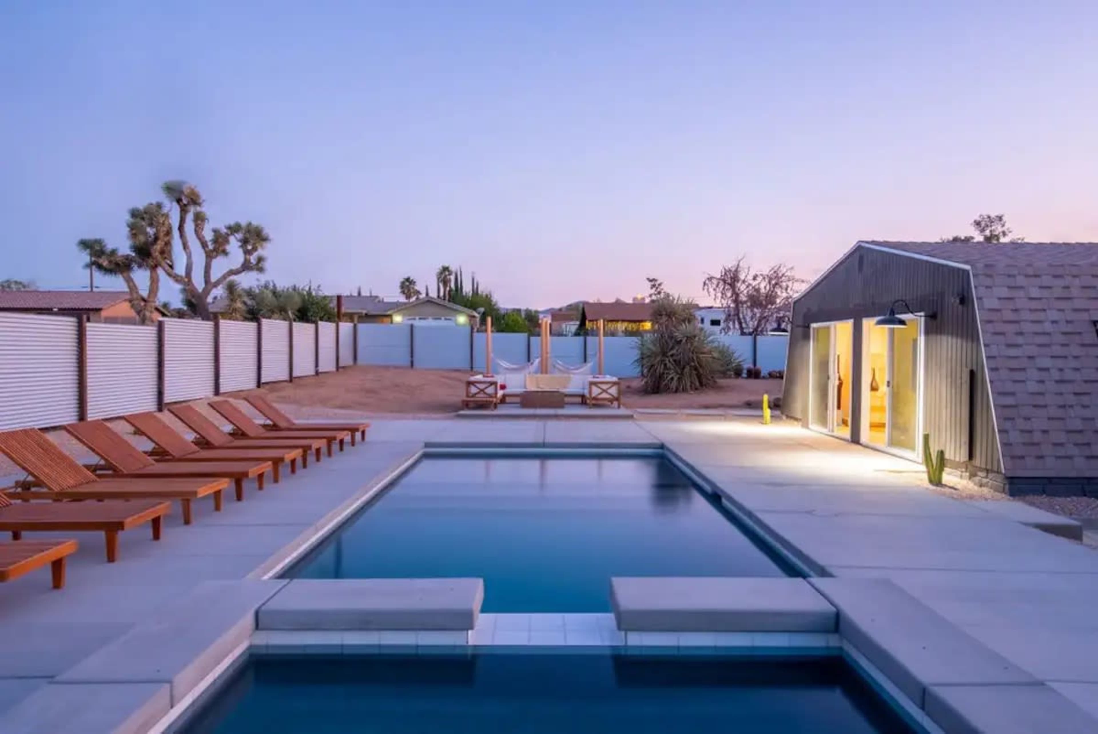A rectangular swimming pool surrounded by wooden lounge chairs, with a modern building in the background and desert vegetation nearby at dusk.