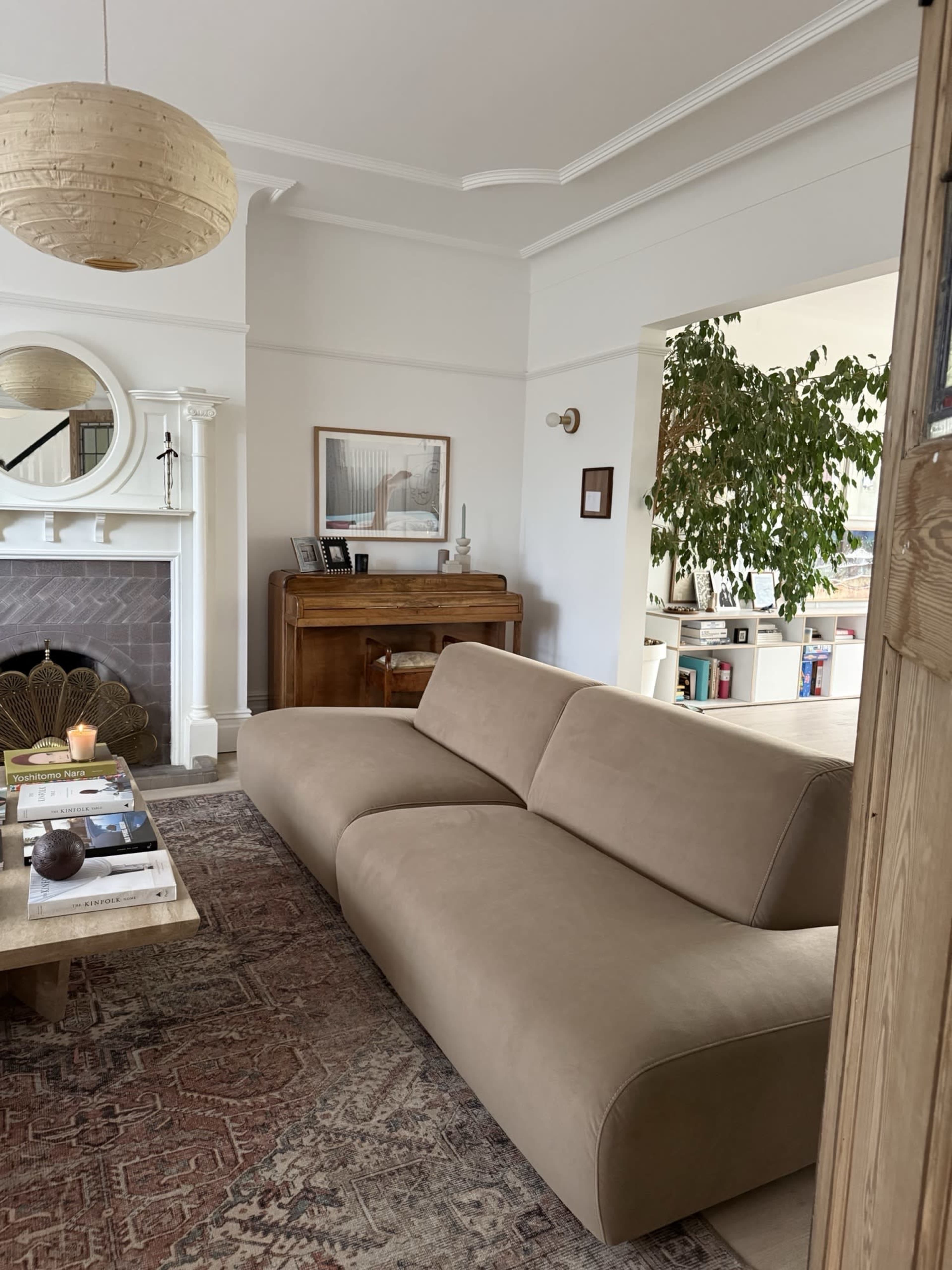 The image shows a modern living room featuring a beige sofa, a wooden piano, a round mirror, and a large indoor plant.
