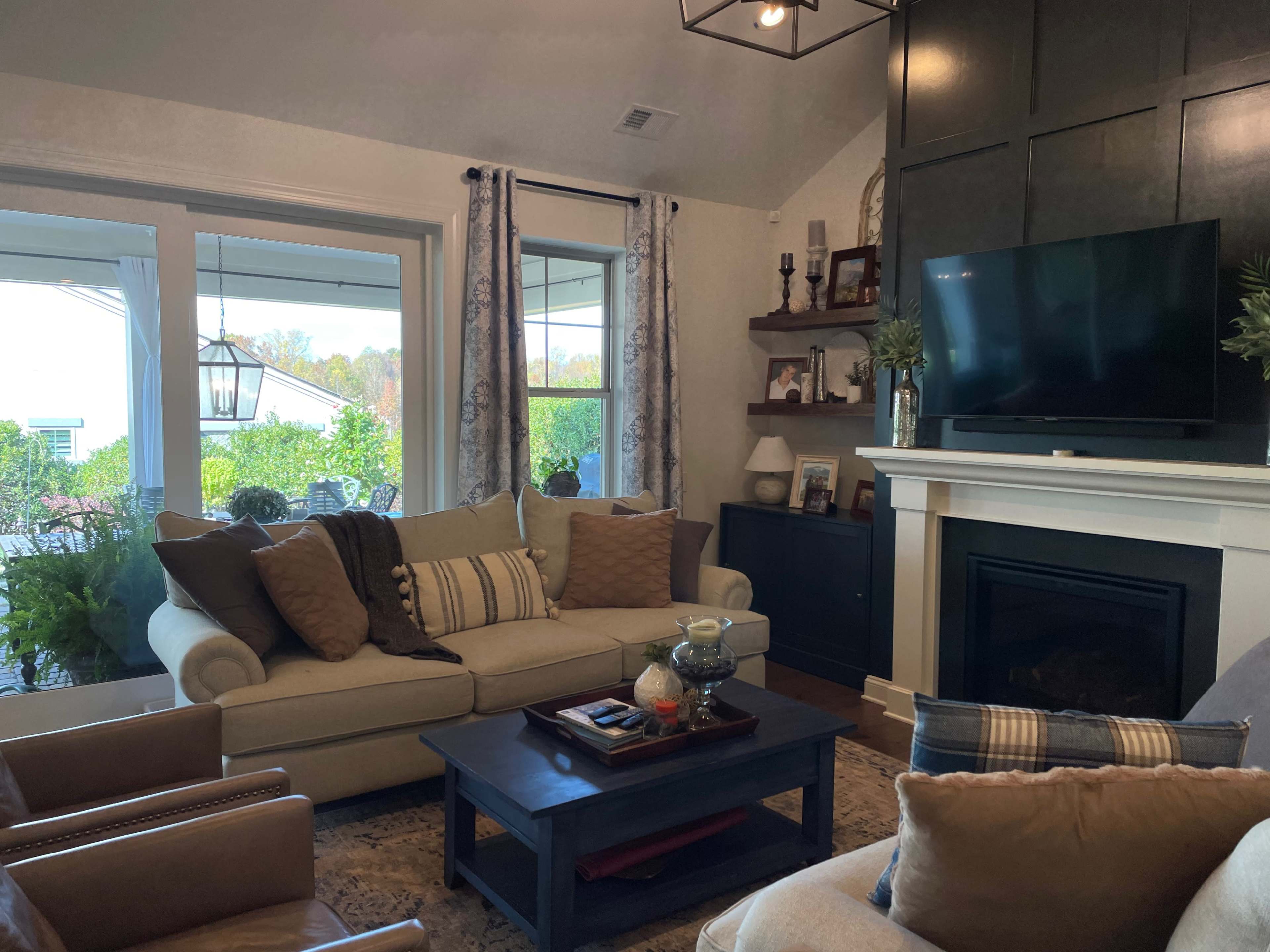A living room features a beige sofa with decorative pillows, a dark coffee table, and a mounted television above a white fireplace, with large windows and curtains framing a view of greenery outside.