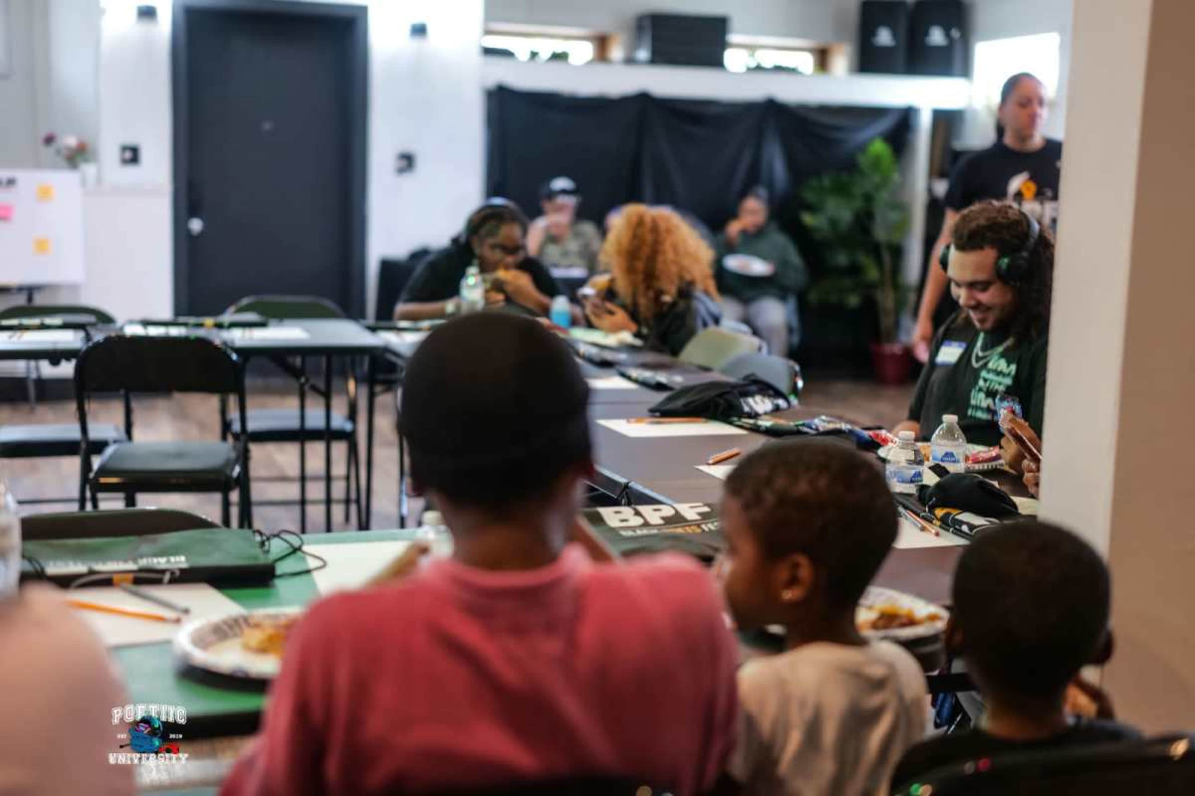 The image shows a group of people seated at tables in a room, some wearing headphones, while others enjoy food and engage with their devices.