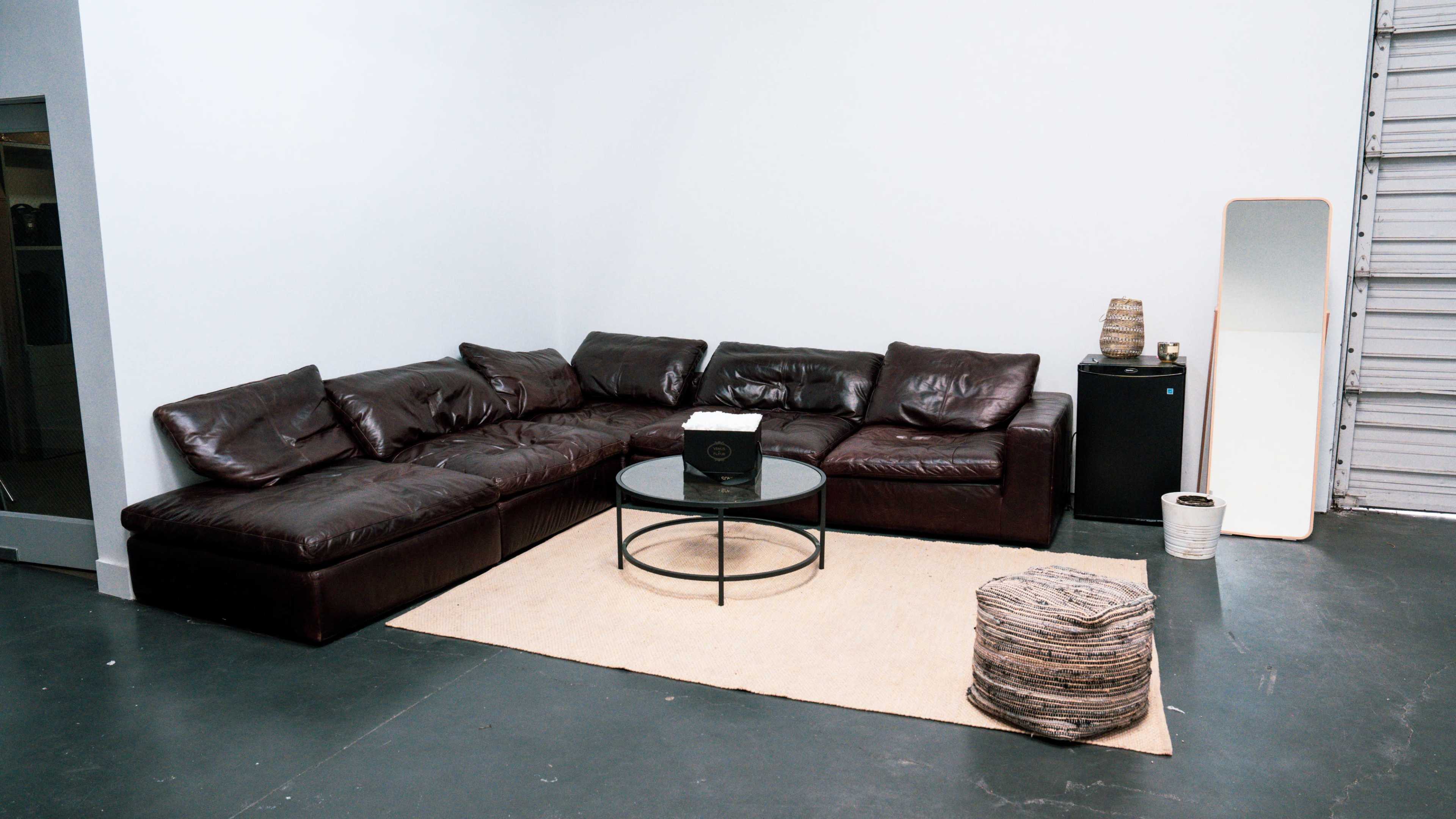 A brown leather sectional sofa is arranged around a circular glass coffee table on a light rug in a minimalist room with a mirror and a small refrigerator.