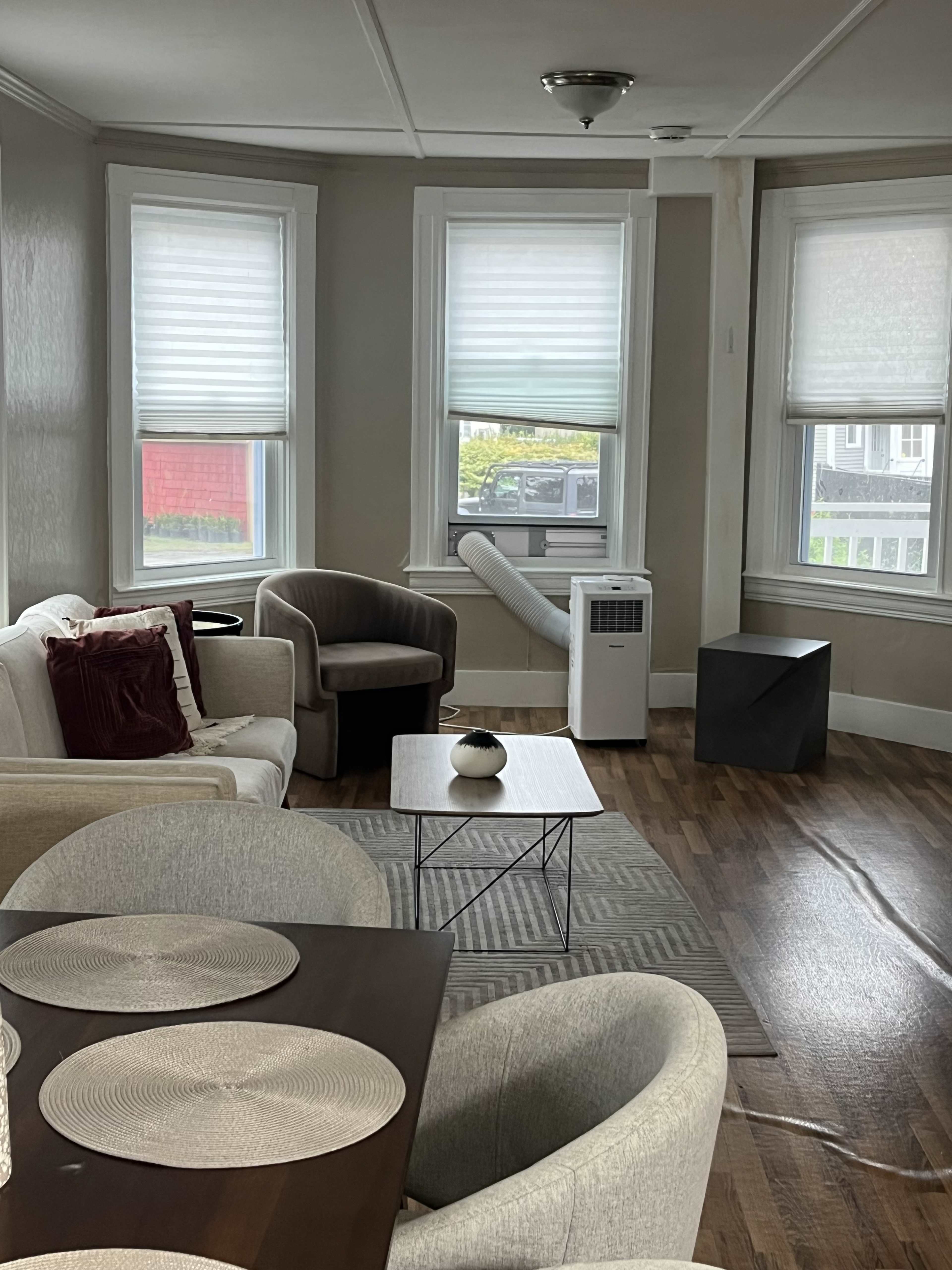 The image shows a living room with two windows, a small dining table, a couch, and an air conditioning unit.