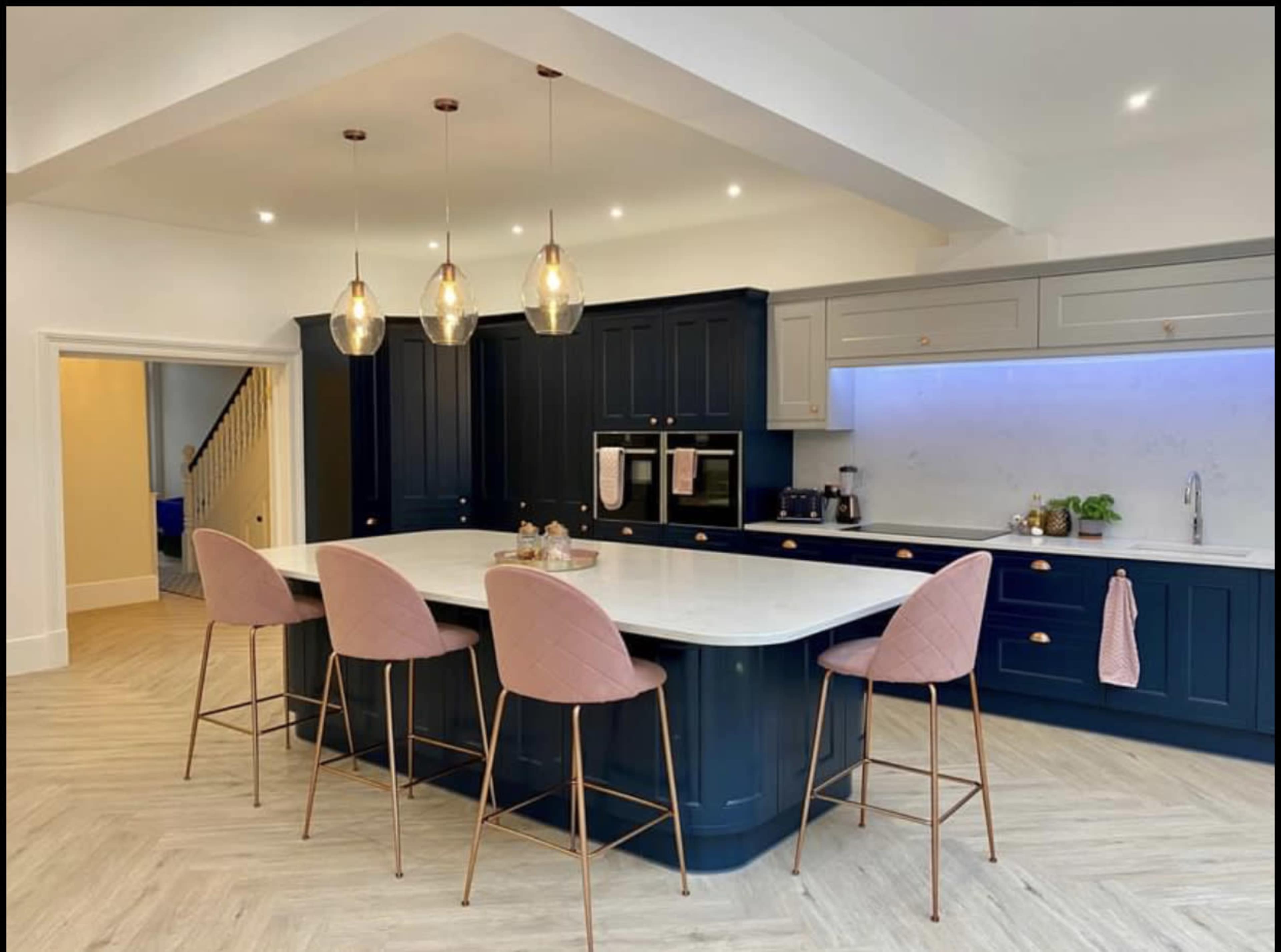 A modern kitchen with a navy blue cabinetry, a white marble island, and four pink upholstered bar stools.