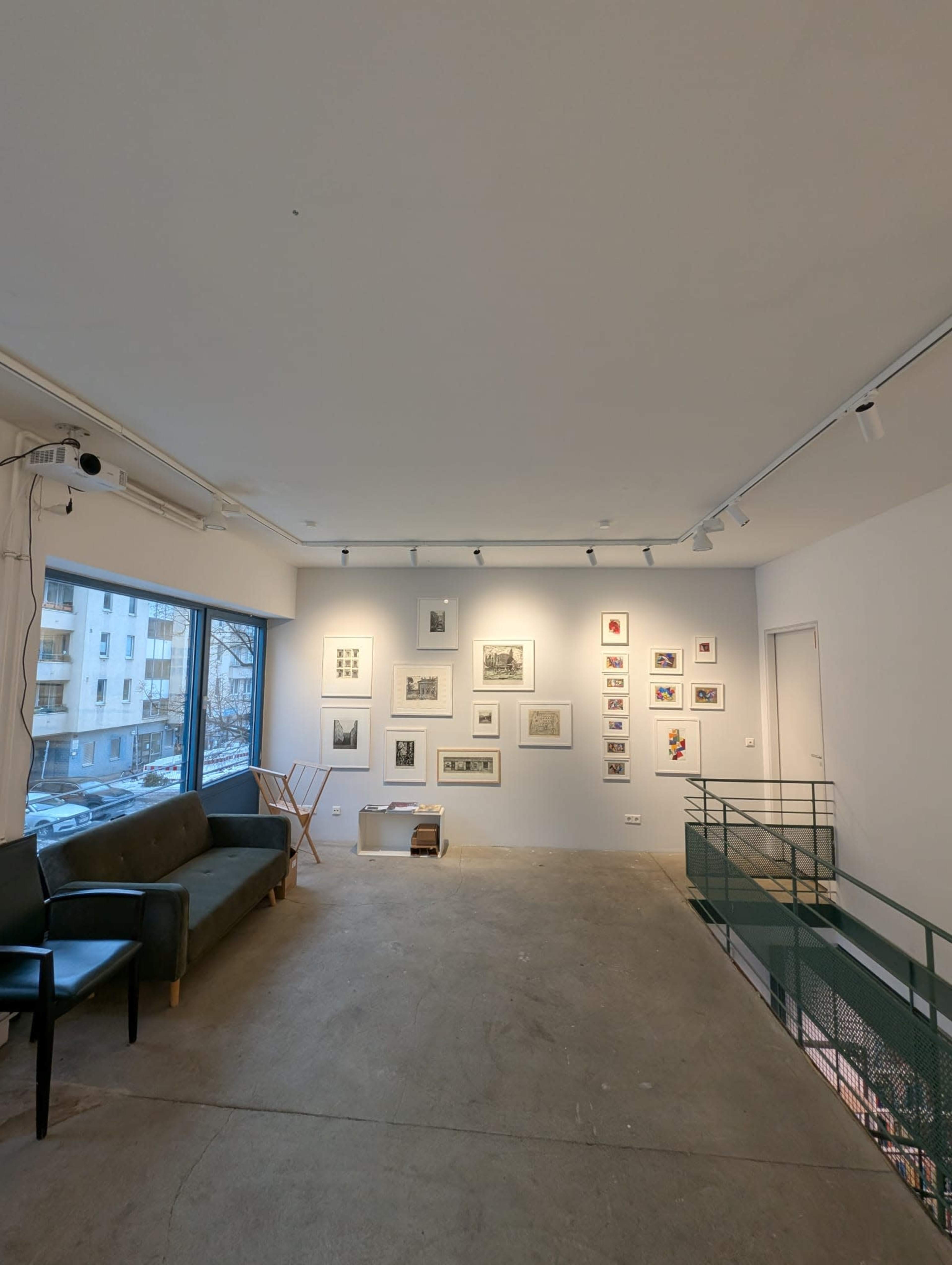 The image shows a spacious art gallery with a green railing, a sofa, and various framed artworks displayed on the walls.