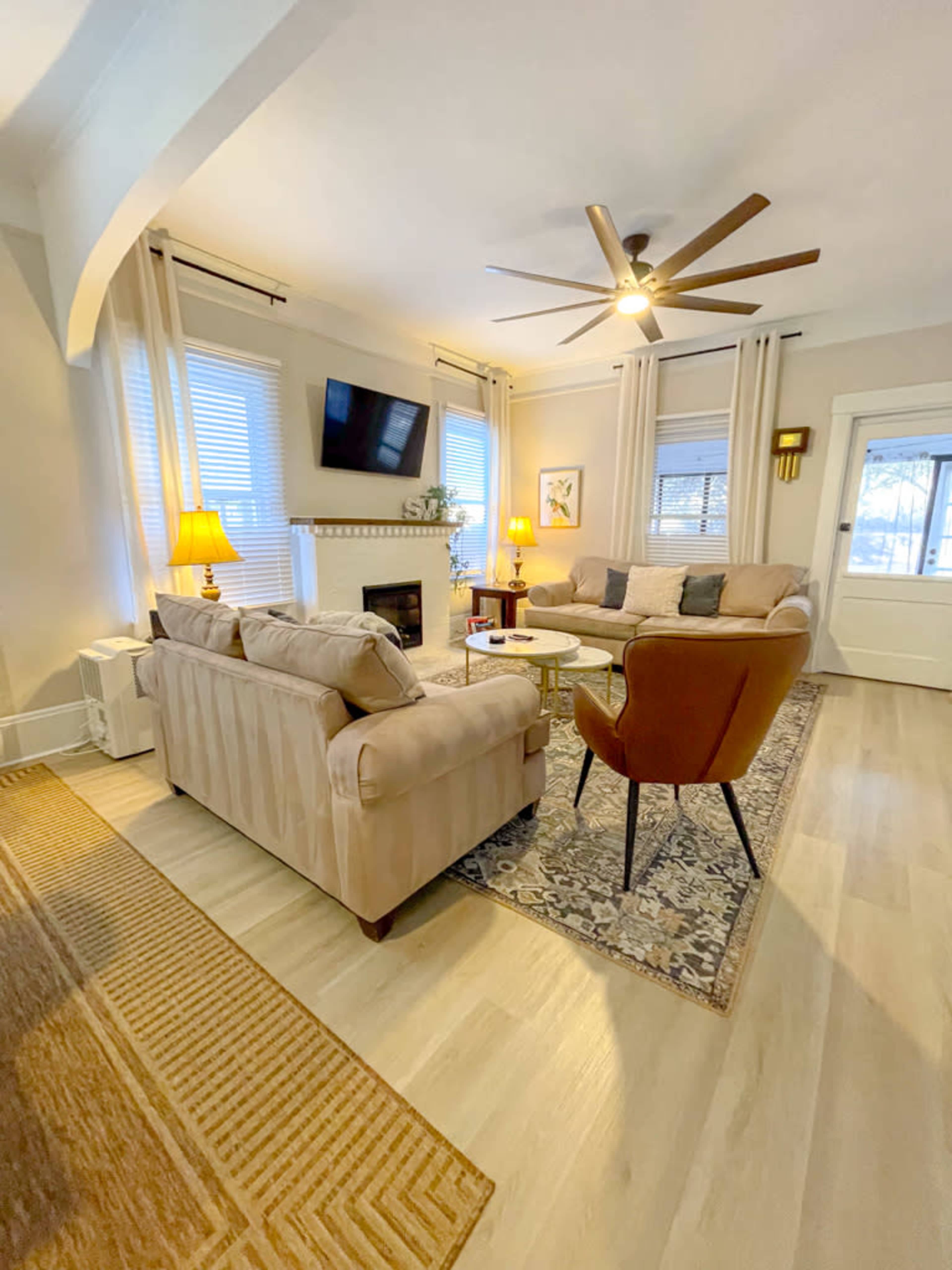 The living room features a beige sectional sofa, a mid-century modern chair, a coffee table, and a wall-mounted television, all illuminated by natural light and lamps.