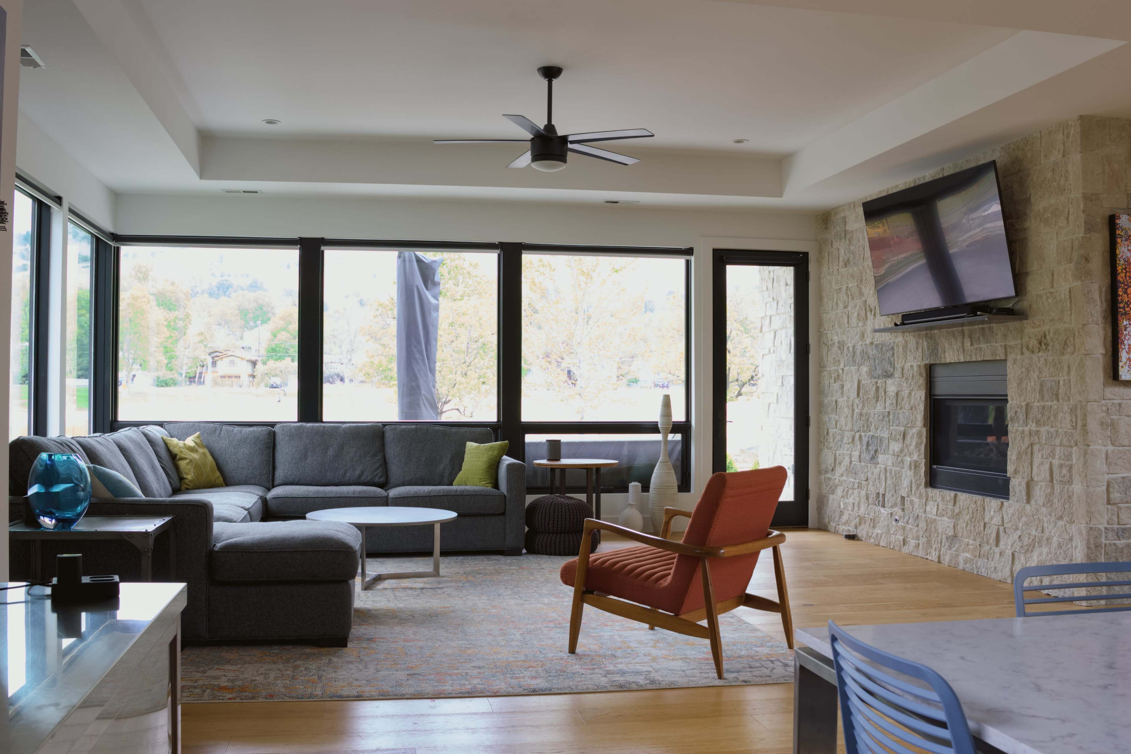 A modern living room features a gray sectional sofa, a round coffee table, and a large window with views of the outdoors, complemented by a stone wall and a fireplace.