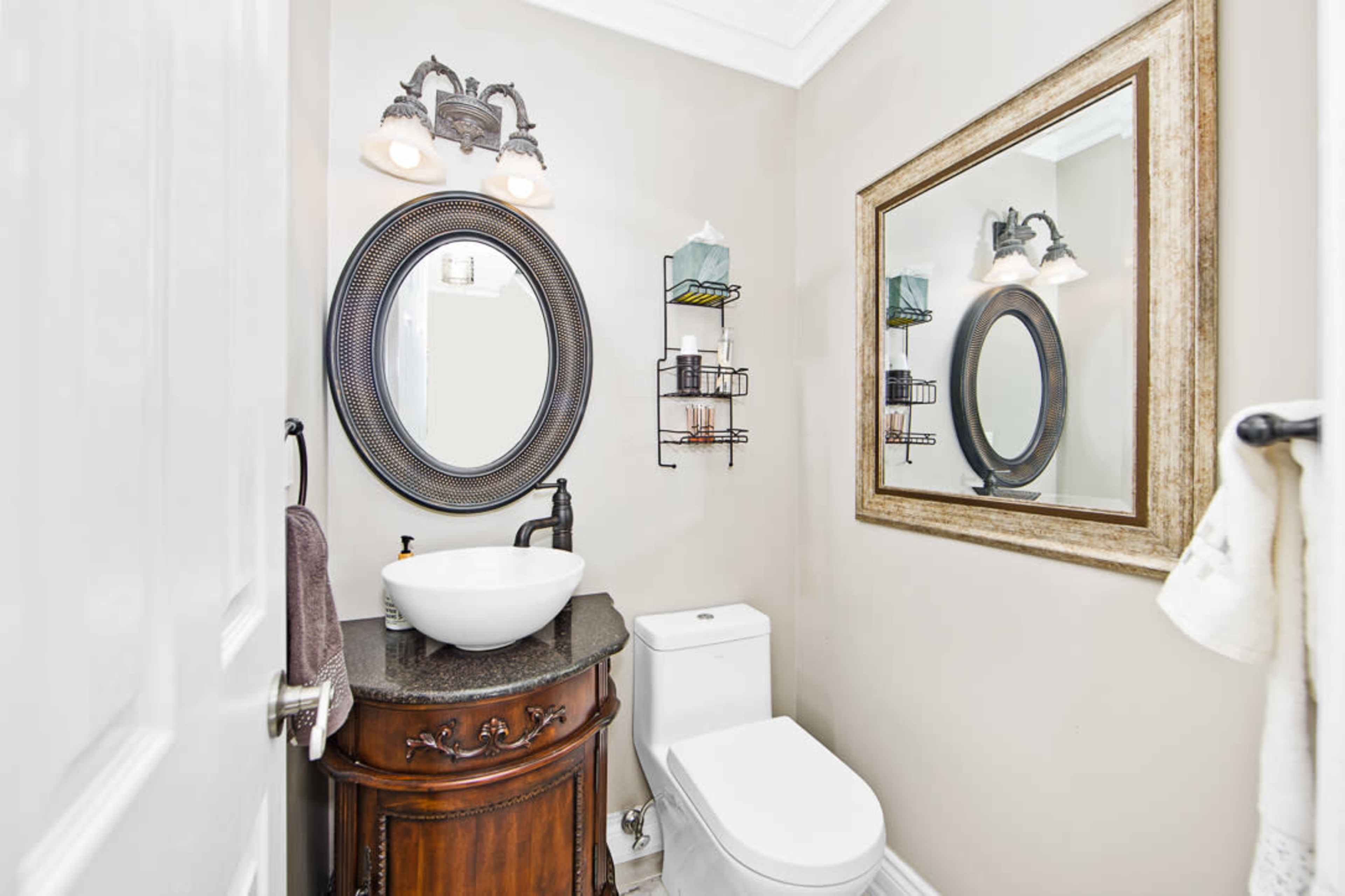 The image shows a small bathroom featuring a round mirror, a wall-mounted storage rack, and a modern sink atop a dark wood vanity.