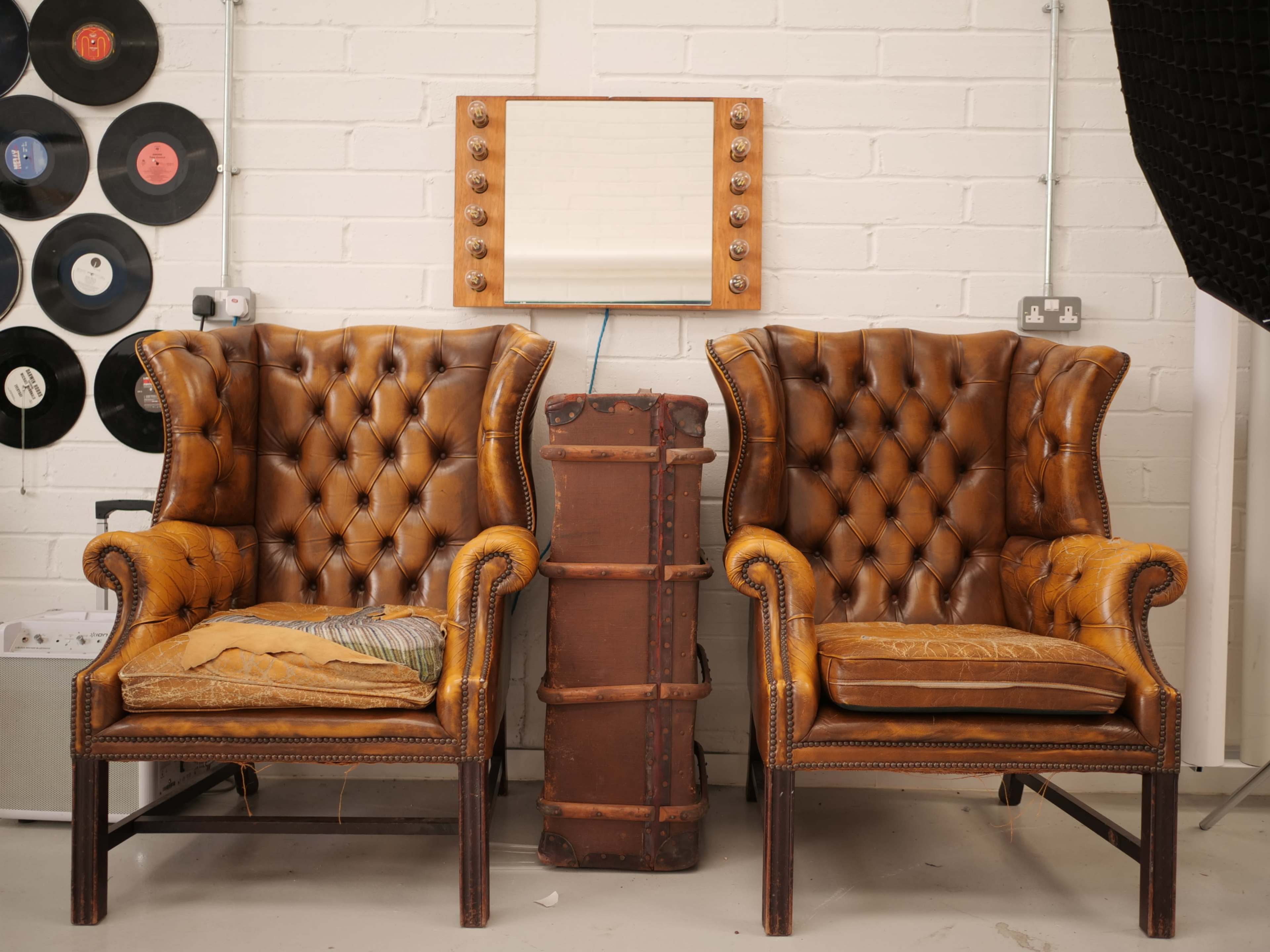 Two vintage leather armchairs are positioned on either side of a suitcase, facing a large illuminated mirror against a white brick wall.
