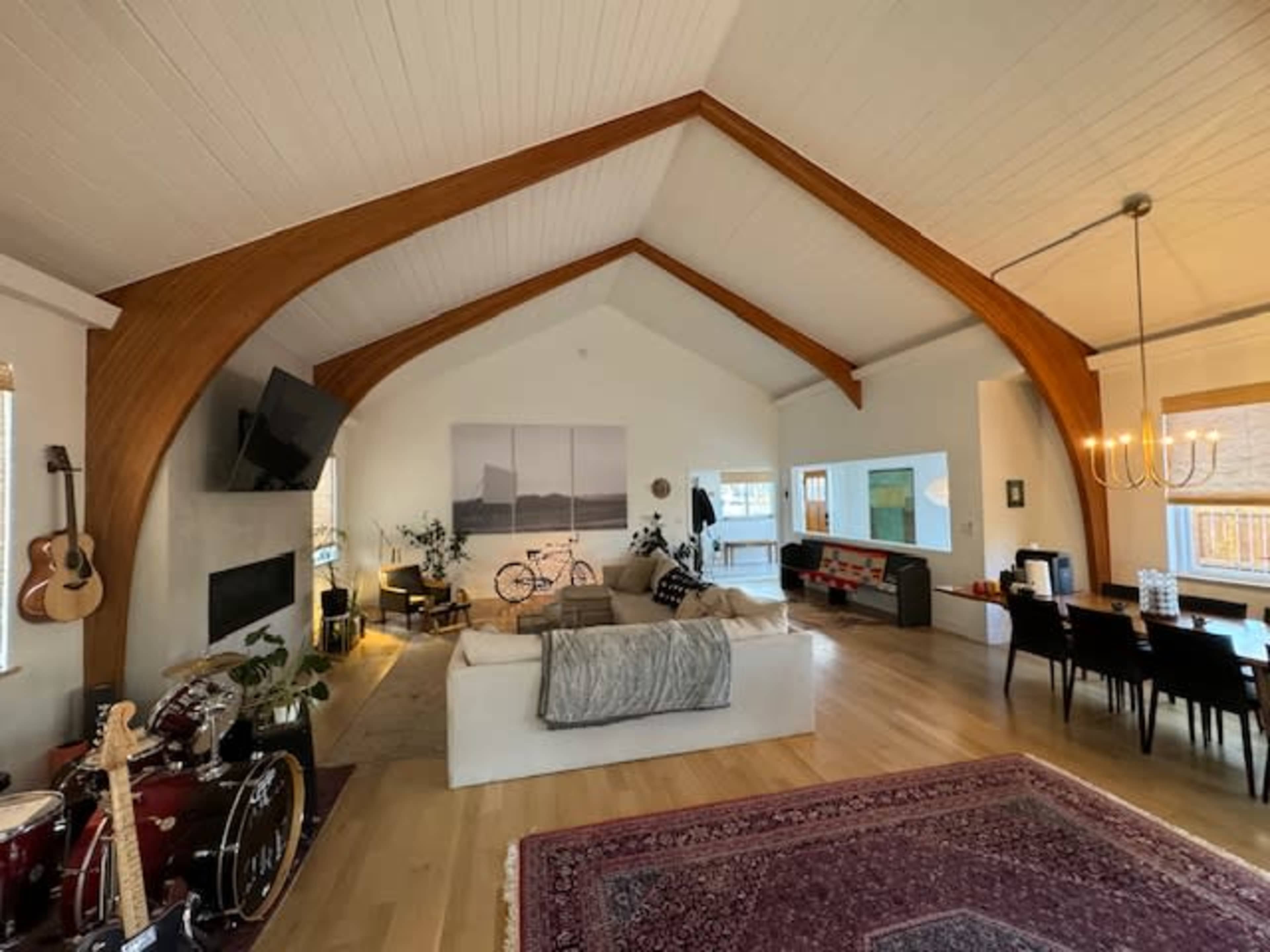 The image shows a spacious living room with a high, vaulted ceiling featuring exposed wooden beams, a large sectional sofa, a dining area, and various decorative elements including musical instruments and plants.