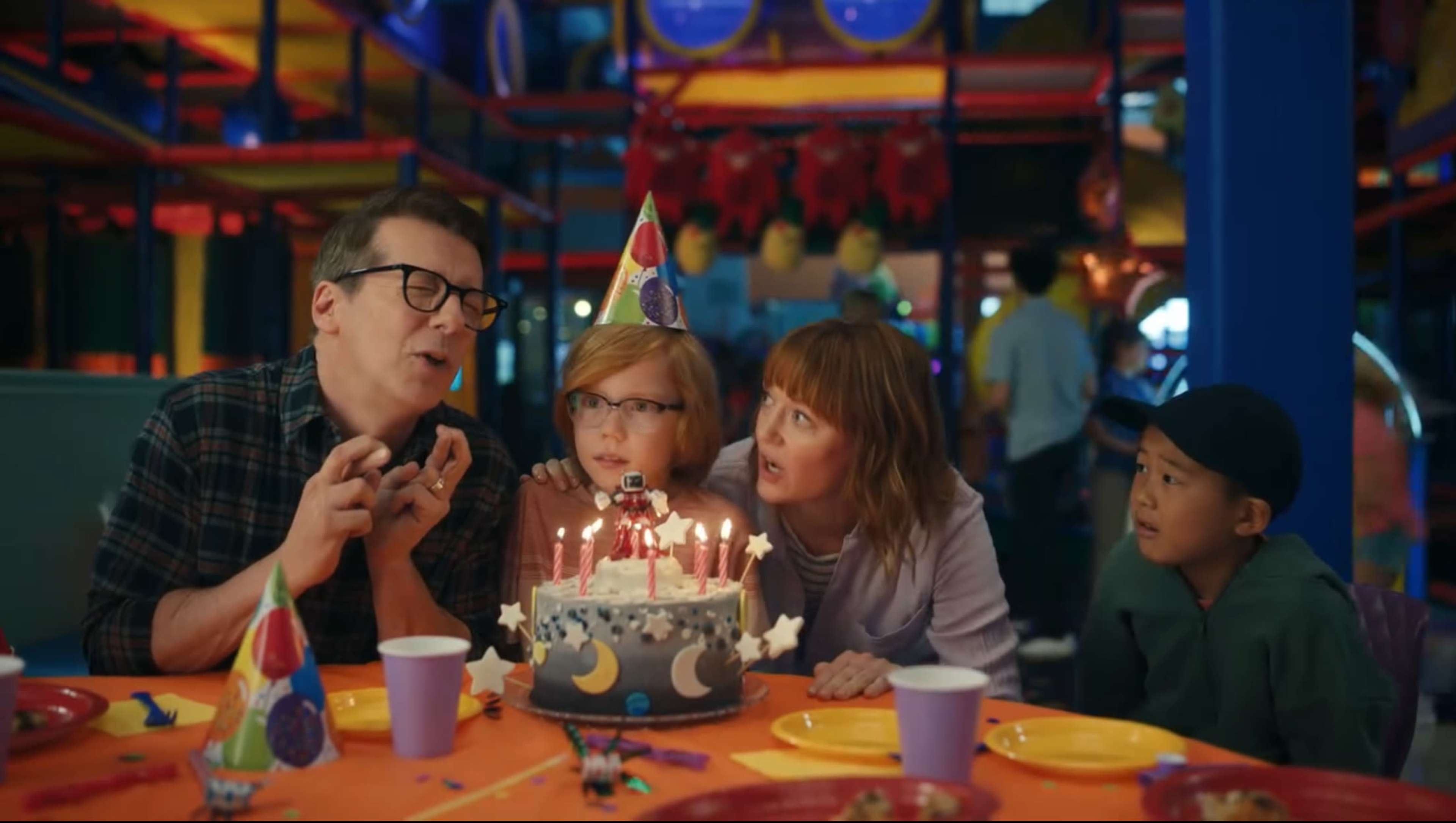 A family gathers around a birthday cake with lit candles while celebrating in a colorful venue decorated for a party.