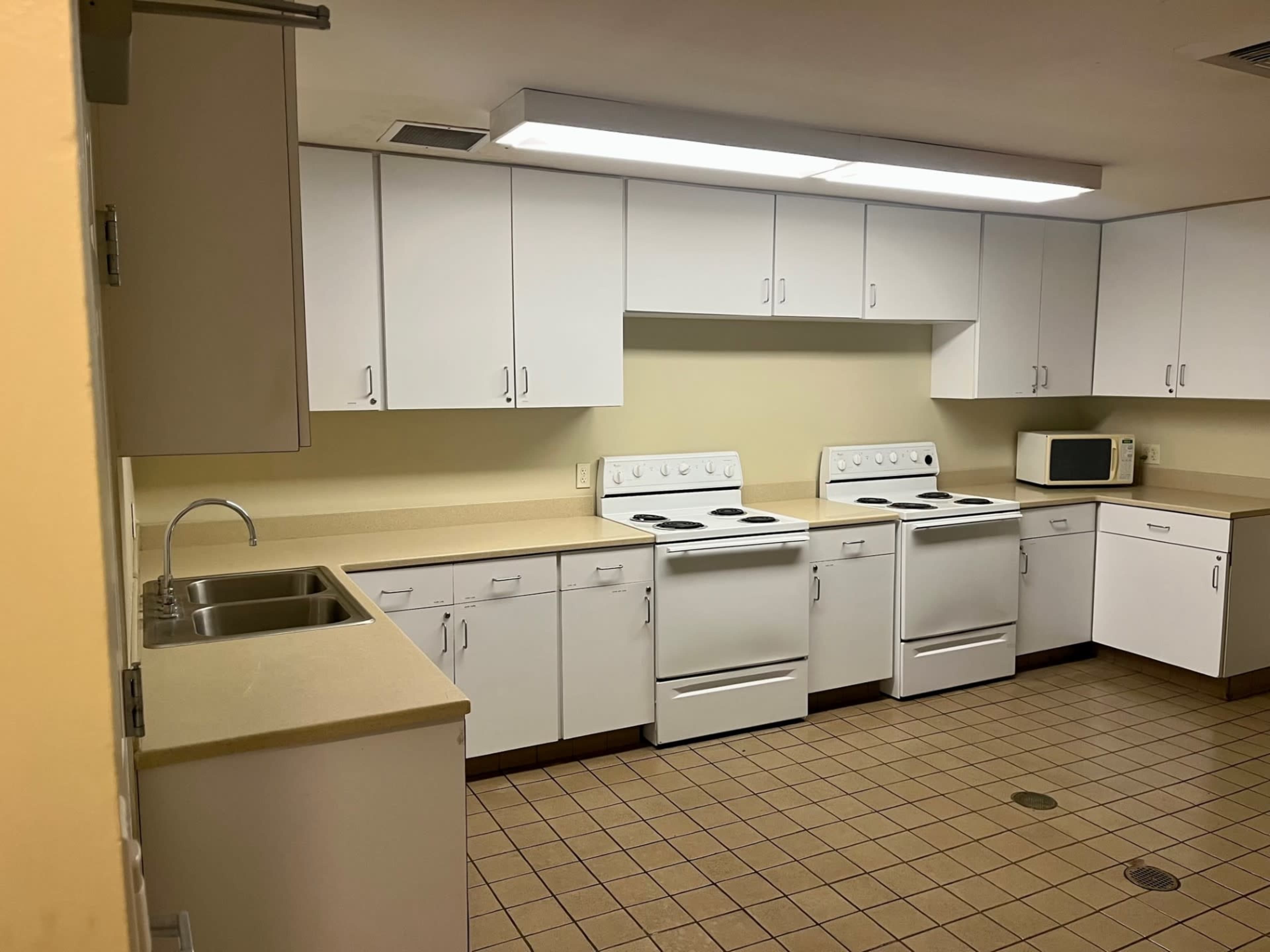 The image shows a kitchen with three white stoves, a sink, a countertop, a microwave, and white cabinets against light-colored walls.