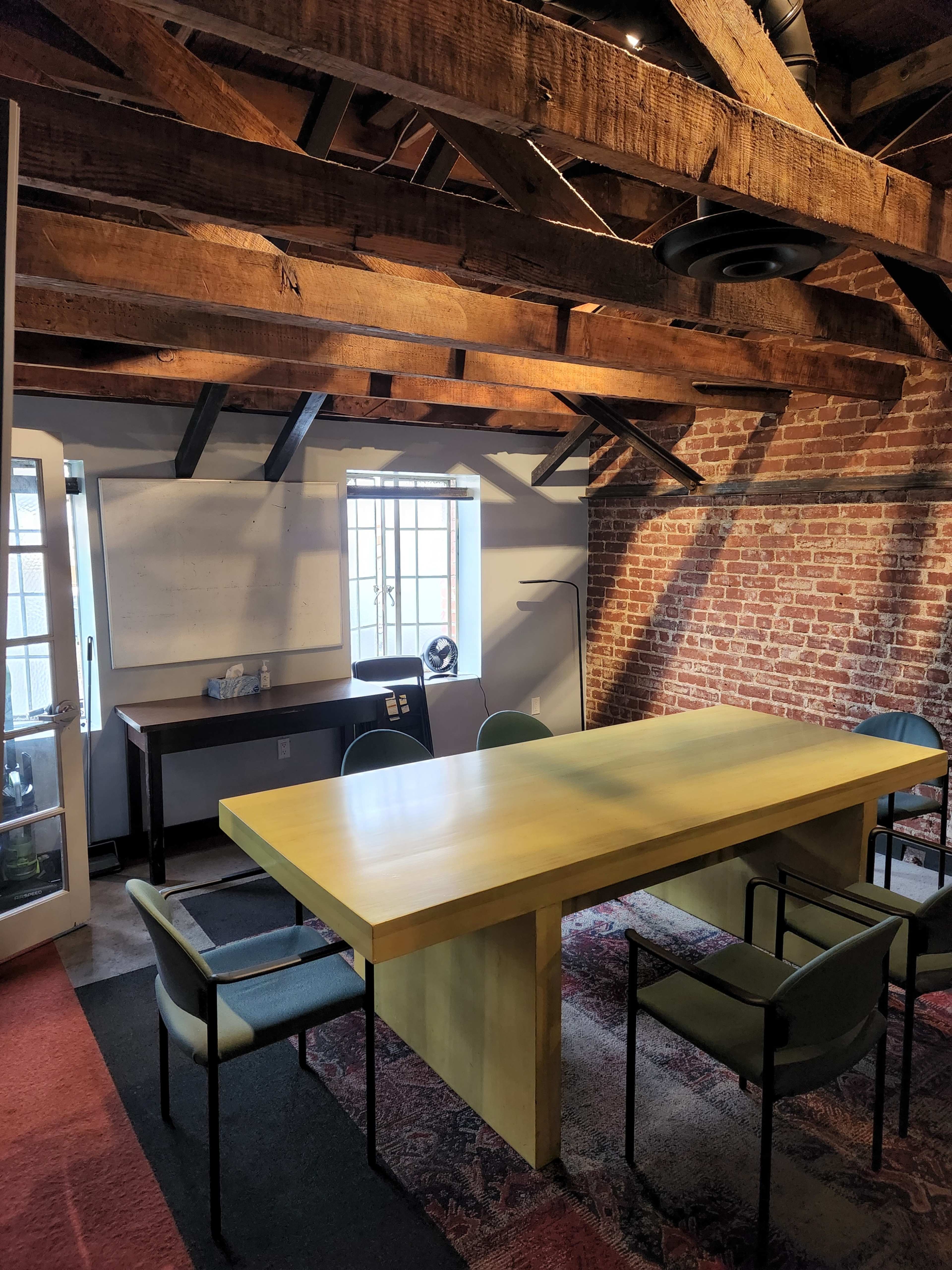 A bright meeting room features a large yellow table surrounded by four green chairs, with exposed wooden beams and a brick wall.