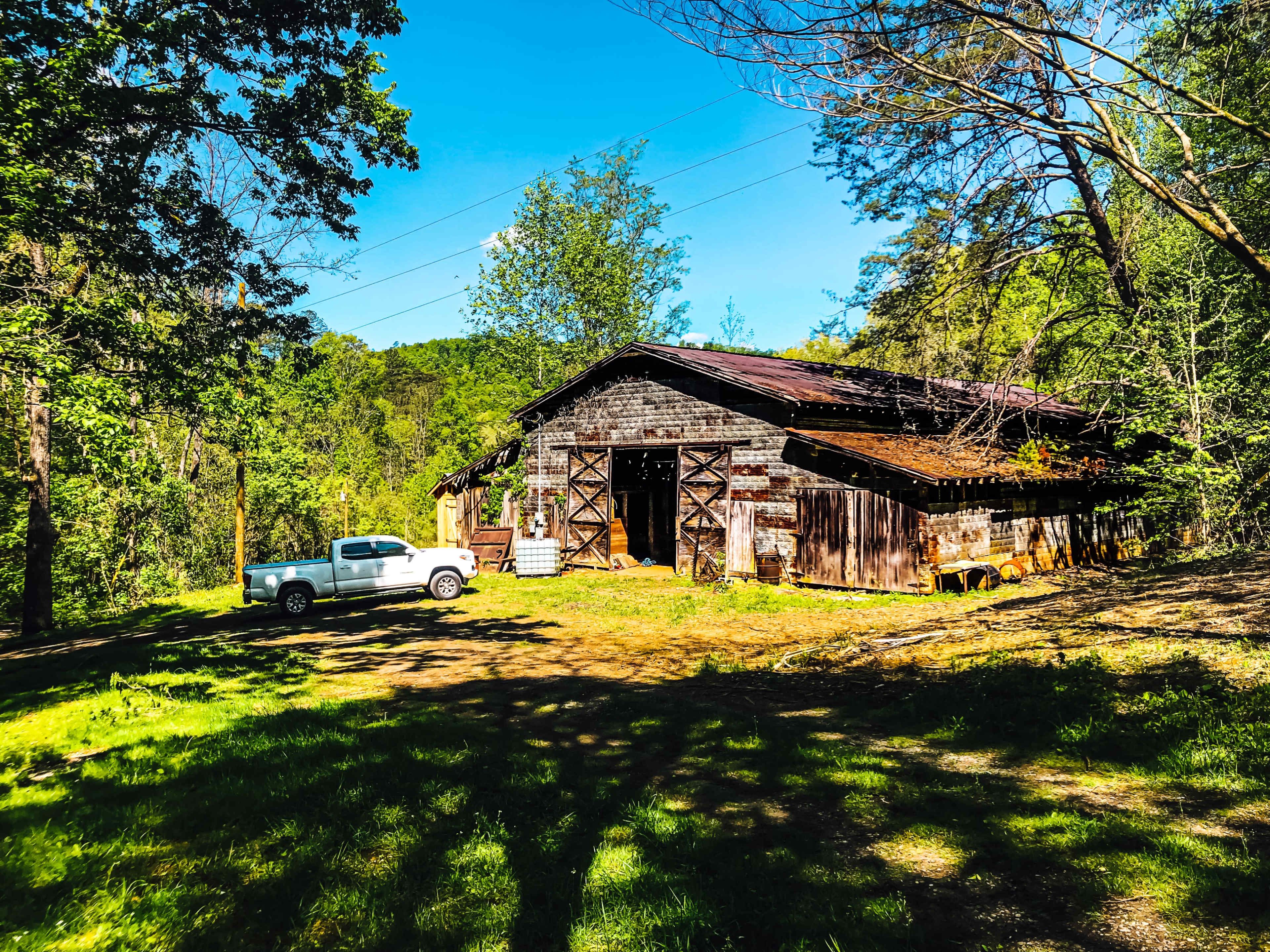 A white pickup truck is parked beside a large, weathered barn surrounded by trees and green grass on a sunny day.