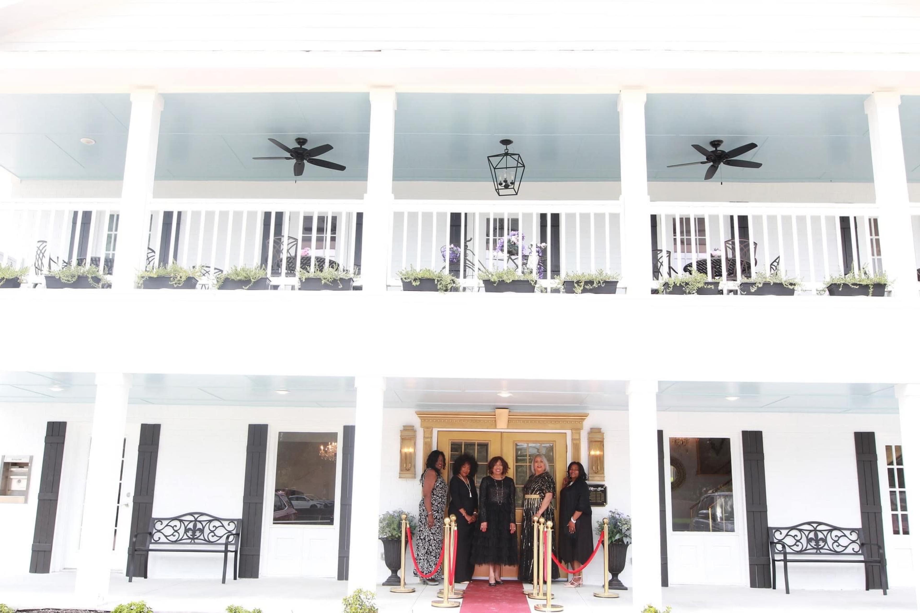 Five women stand in front of a white-featured building with a red carpet and gold stanchions.