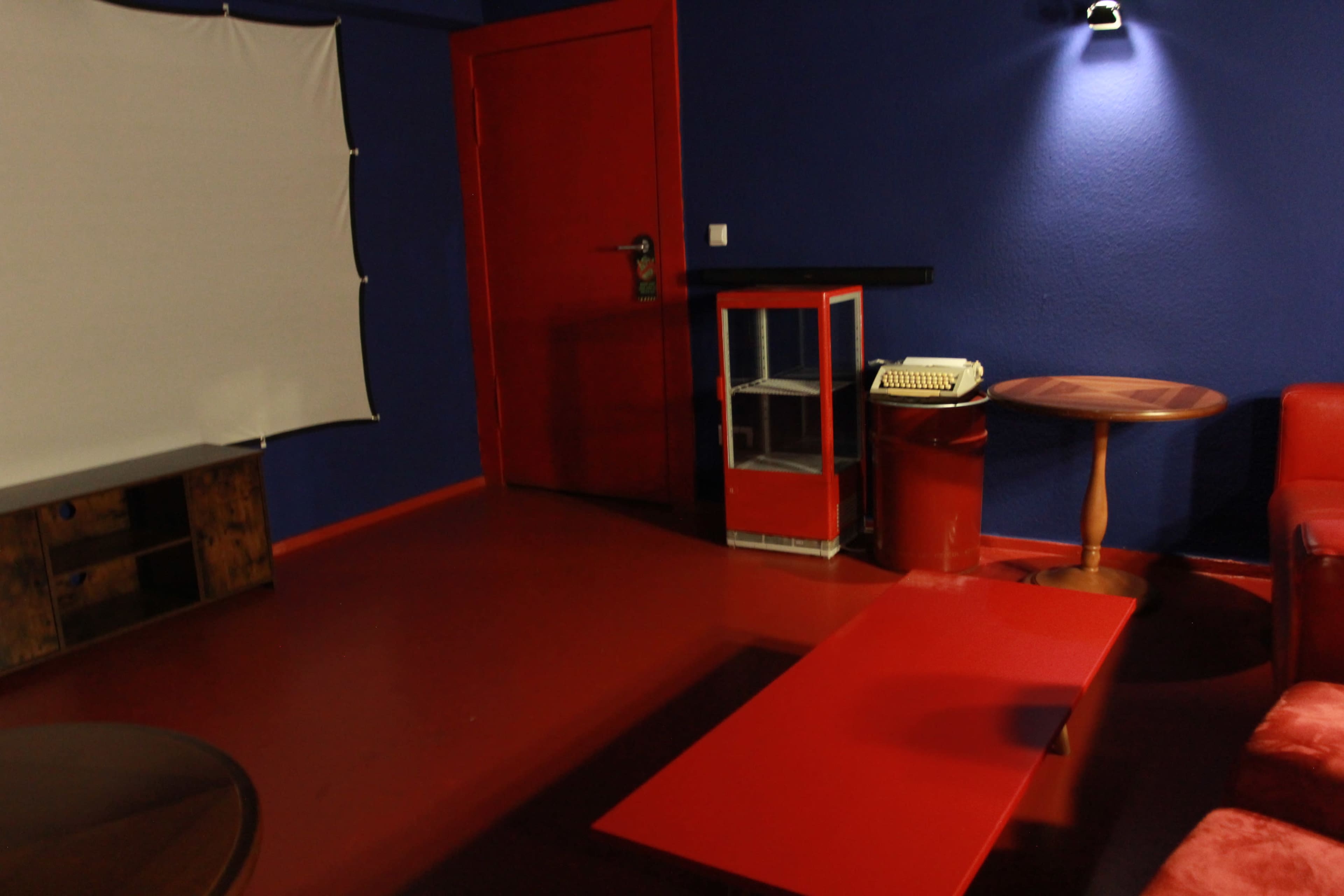 The image shows a small room with blue walls, a red door, a white projection screen, a table, a typewriter, and red furniture.