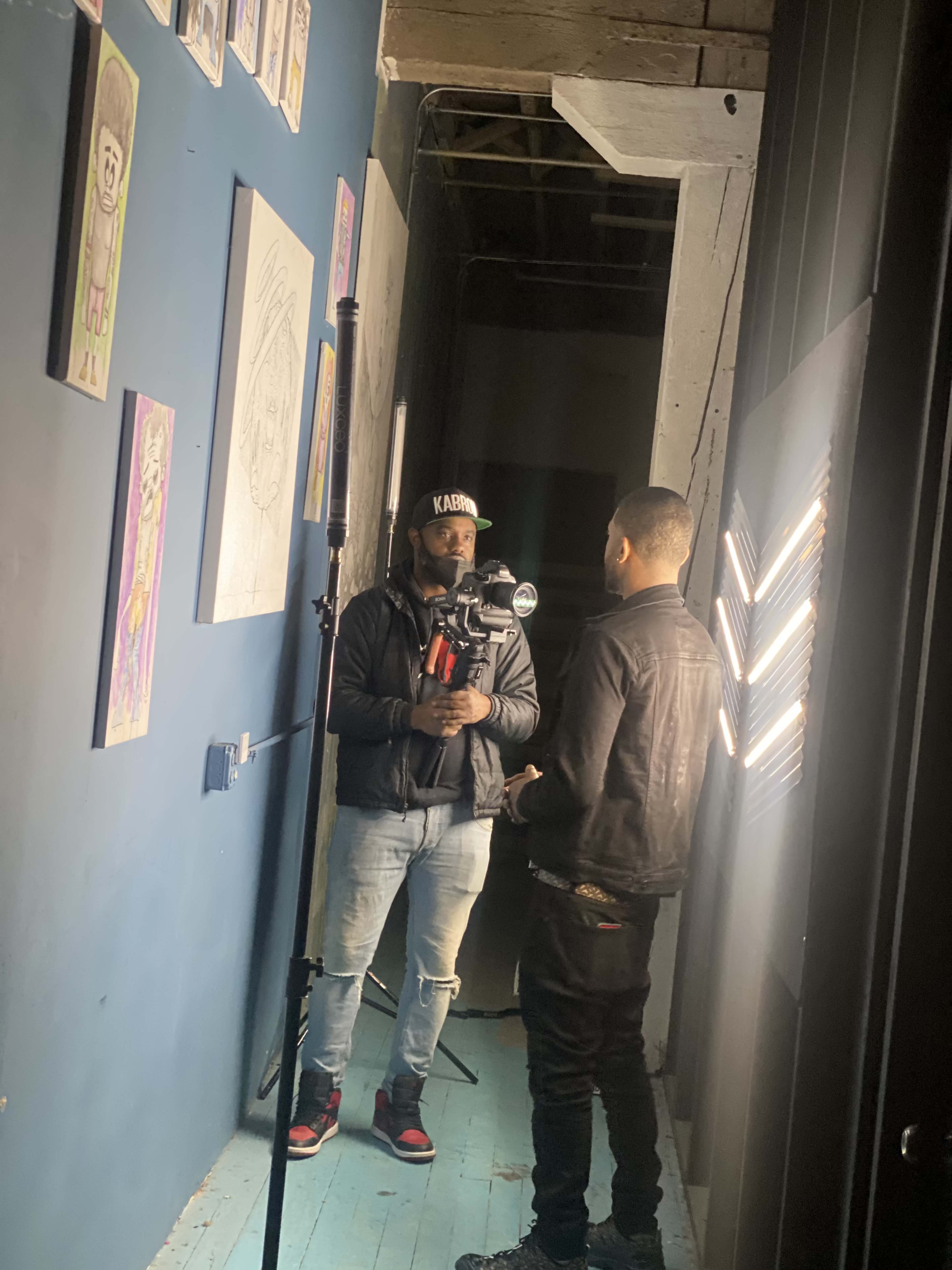 Two men are standing in a narrow hallway with artwork on the walls, one holding a camera and the other engaged in conversation.