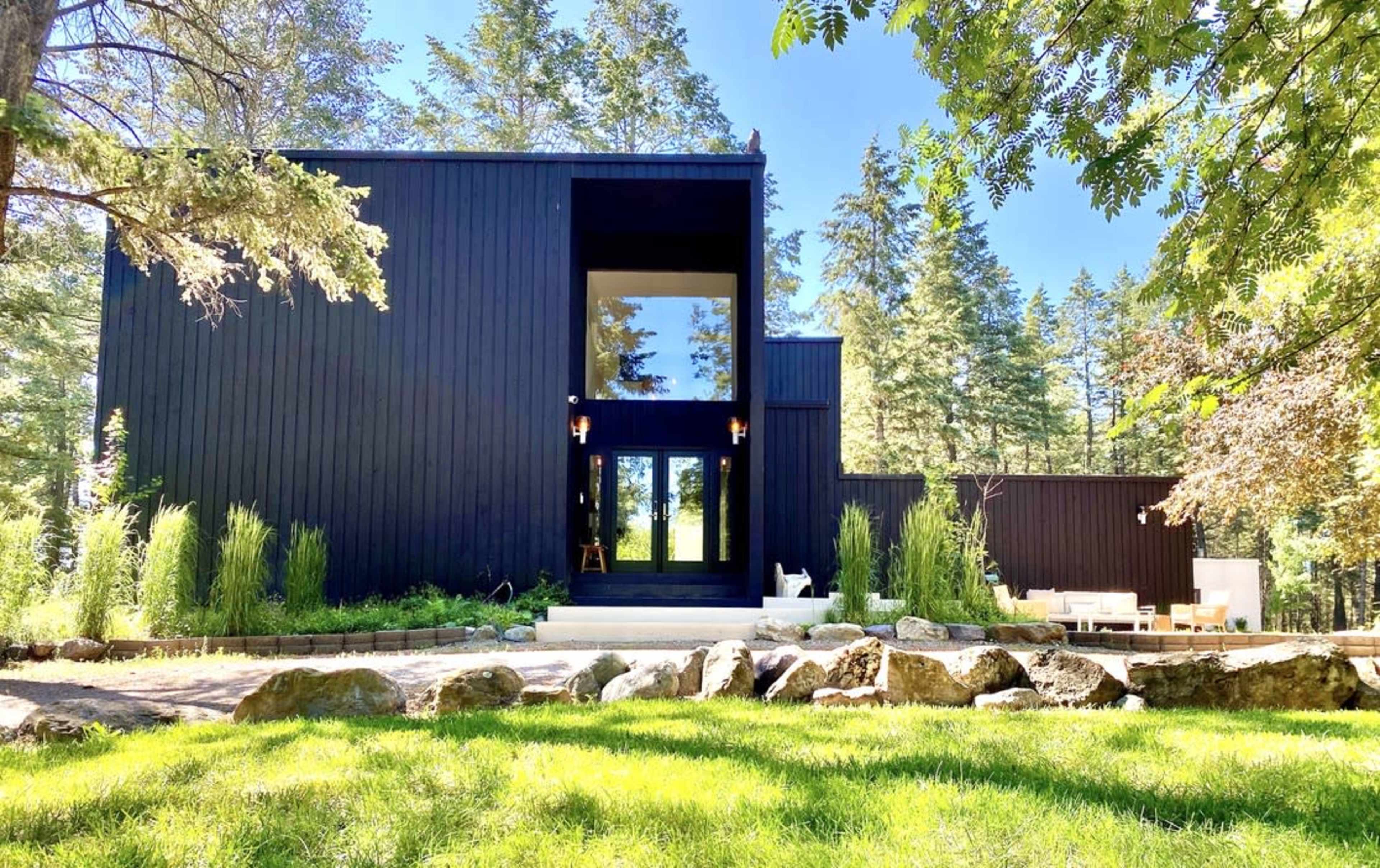 A modern black house with large windows is surrounded by green grass, trees, and landscaped rocks.
