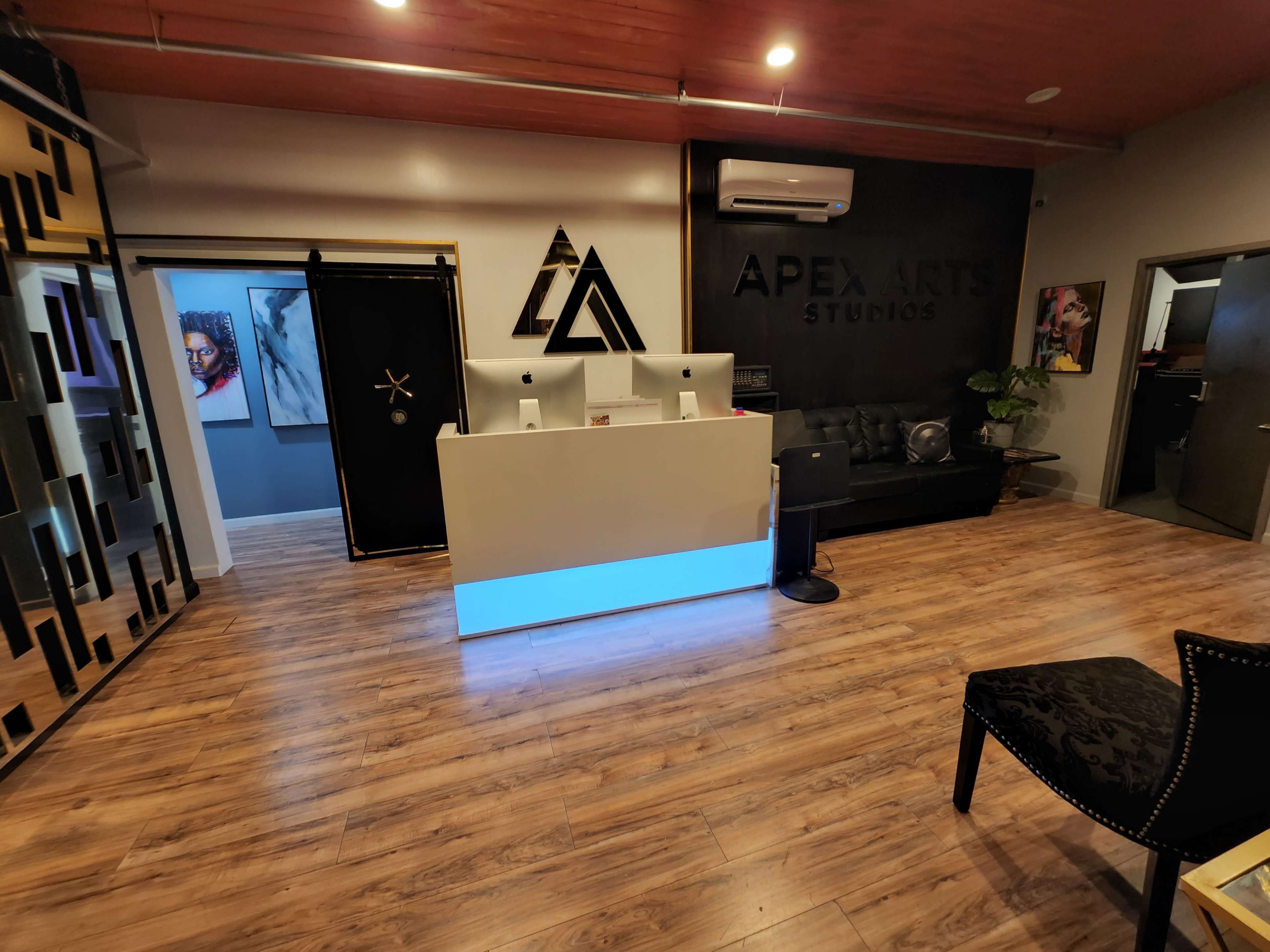 The image shows a modern reception area with a sleek desk illuminated from below, featuring computer displays, and an artistic logo on the wall beside a seating area.