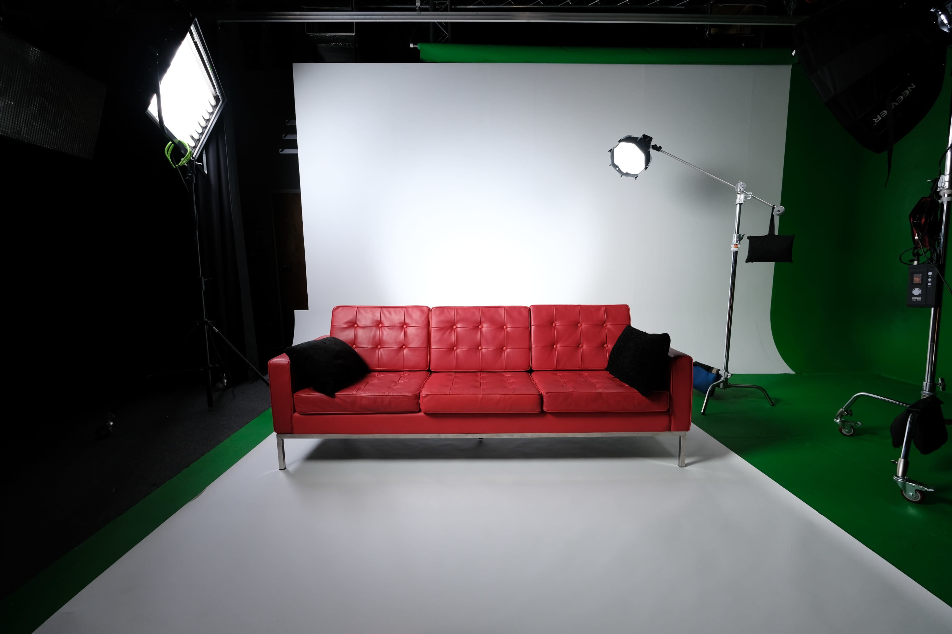 A red sofa with black cushions is positioned in front of a gray backdrop and green screen in a studio setting.