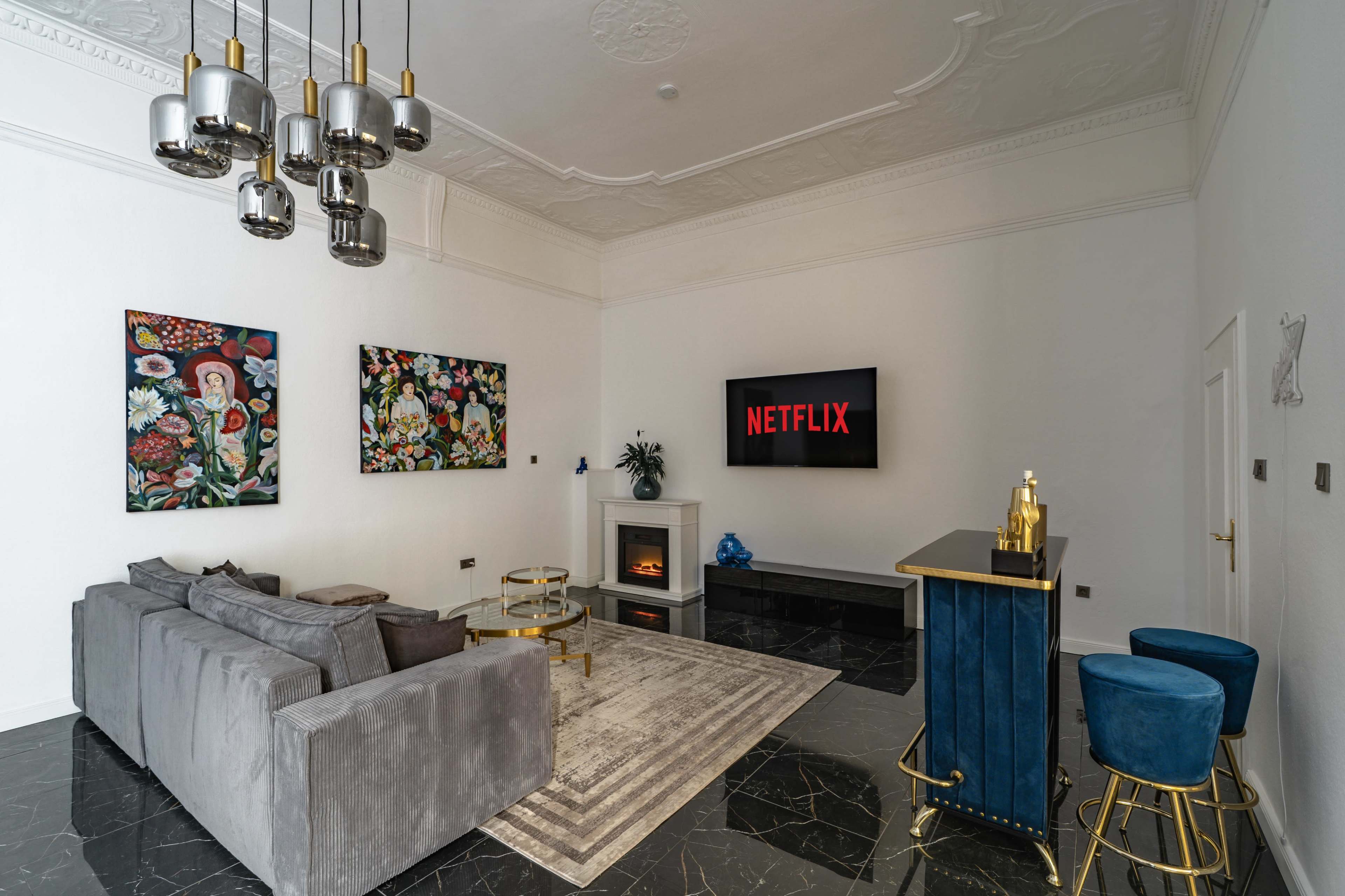 The image depicts a modern living room featuring a gray sectional sofa, two blue bar stools, a flat-screen TV displaying the Netflix logo, and artistic wall decorations.