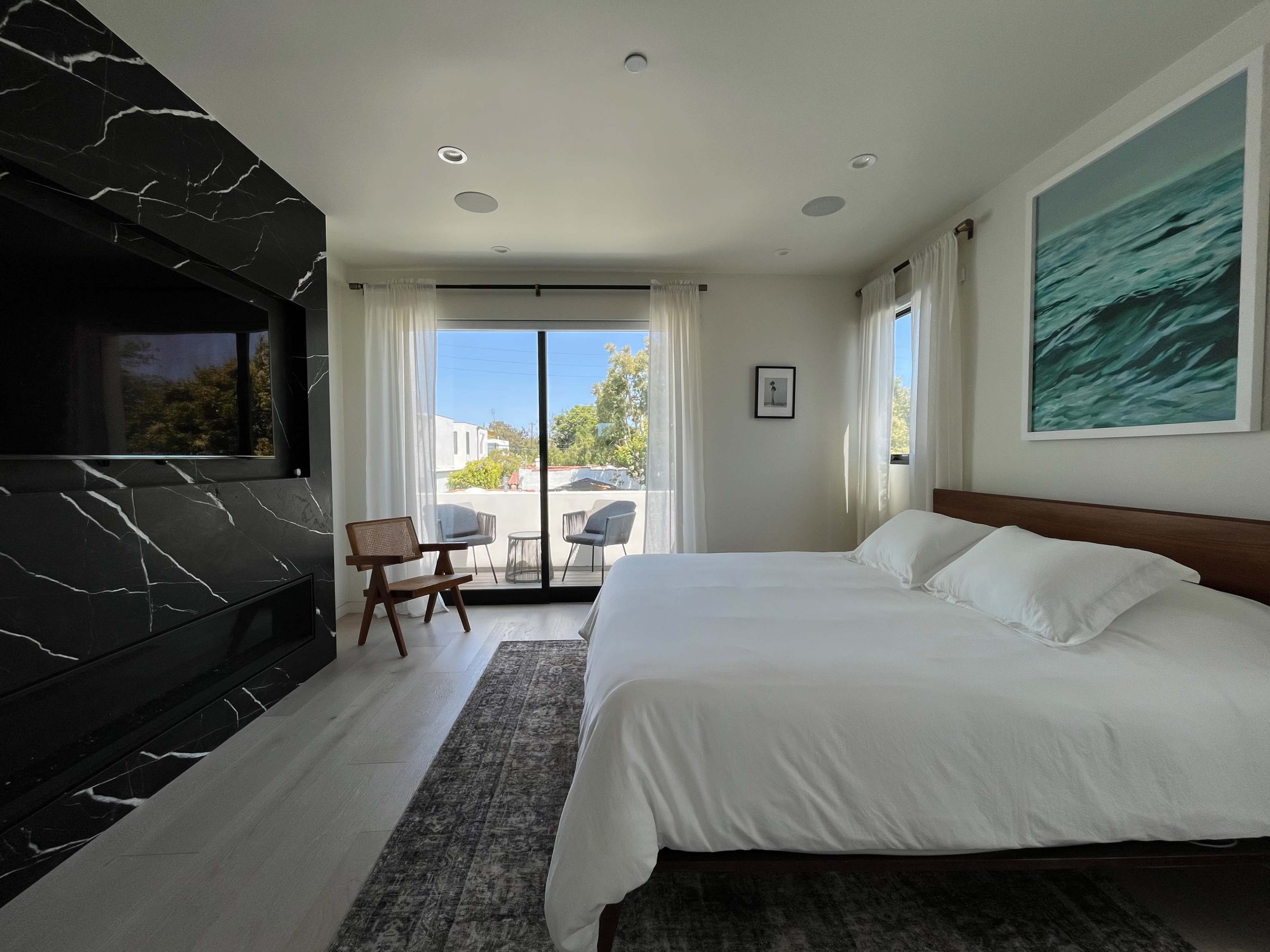 The image shows a modern bedroom featuring a large bed, a black marble accent wall with a television, and a balcony visible through glass doors.