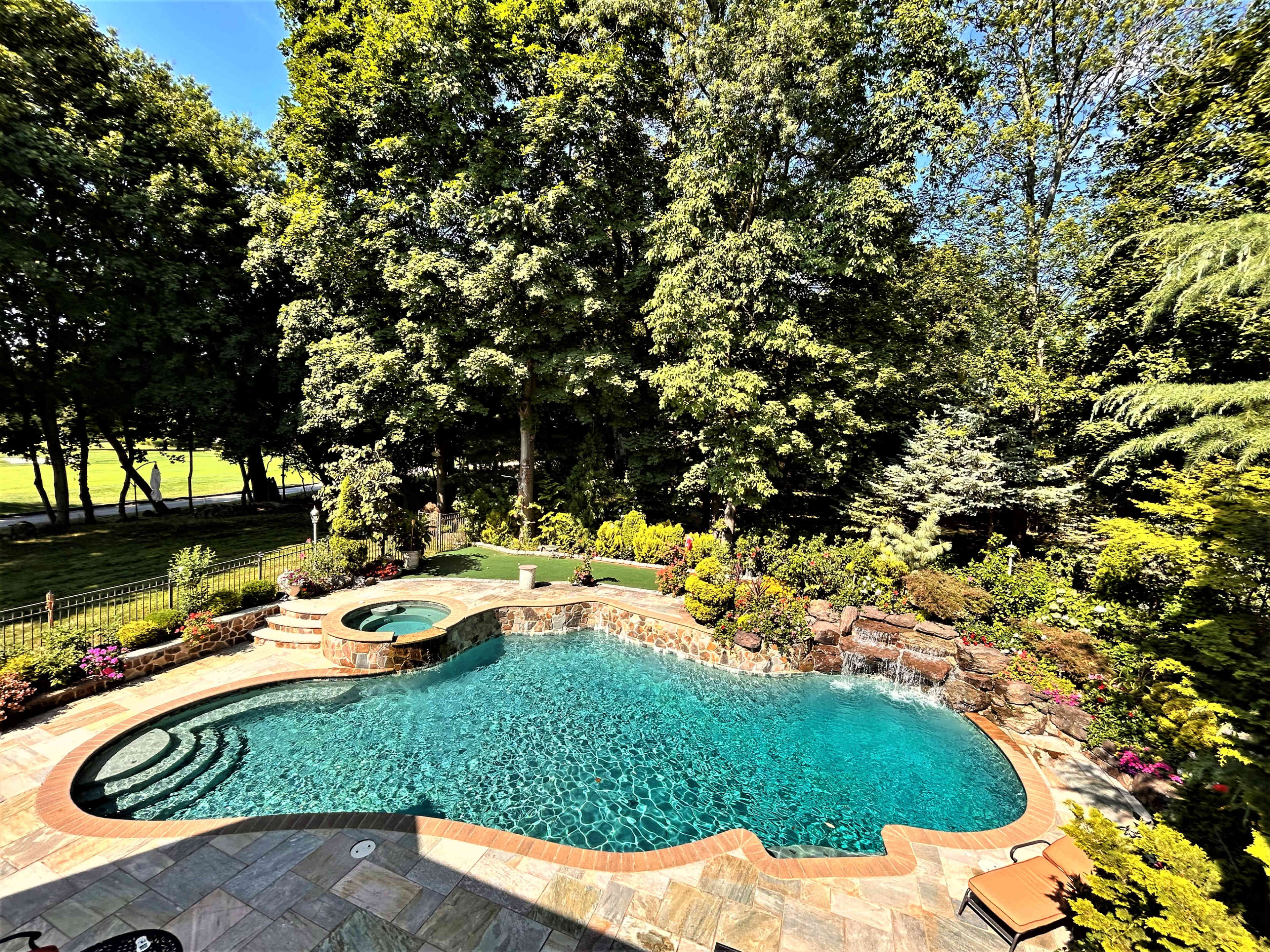 The image shows a large, landscaped backyard swimming pool with a spa, surrounded by trees and vibrant greenery.