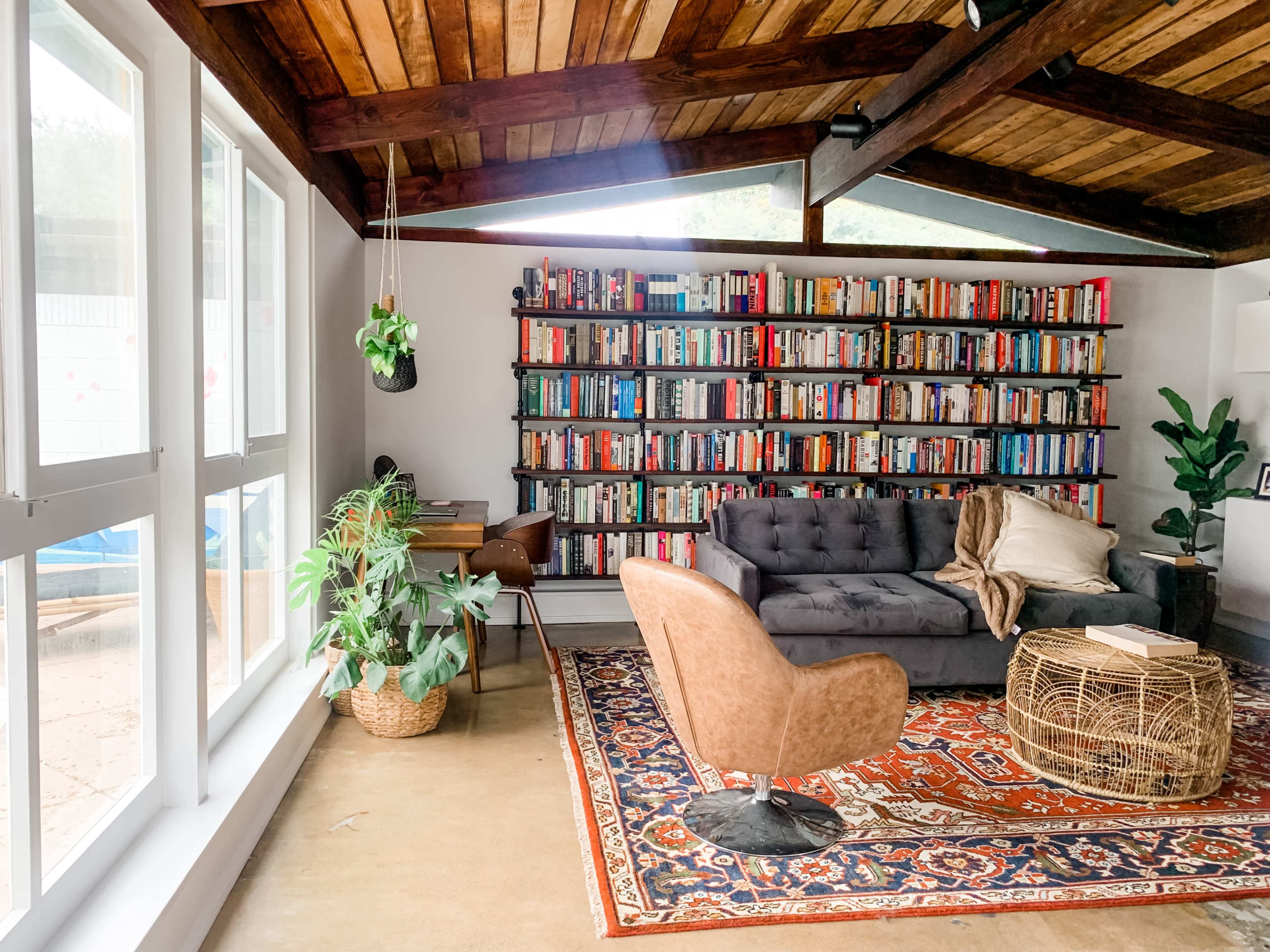 A cozy living room features a large bookshelf filled with colorful books, a plush sofa, a stylish armchair, and potted plants, with ample natural light coming through the windows.