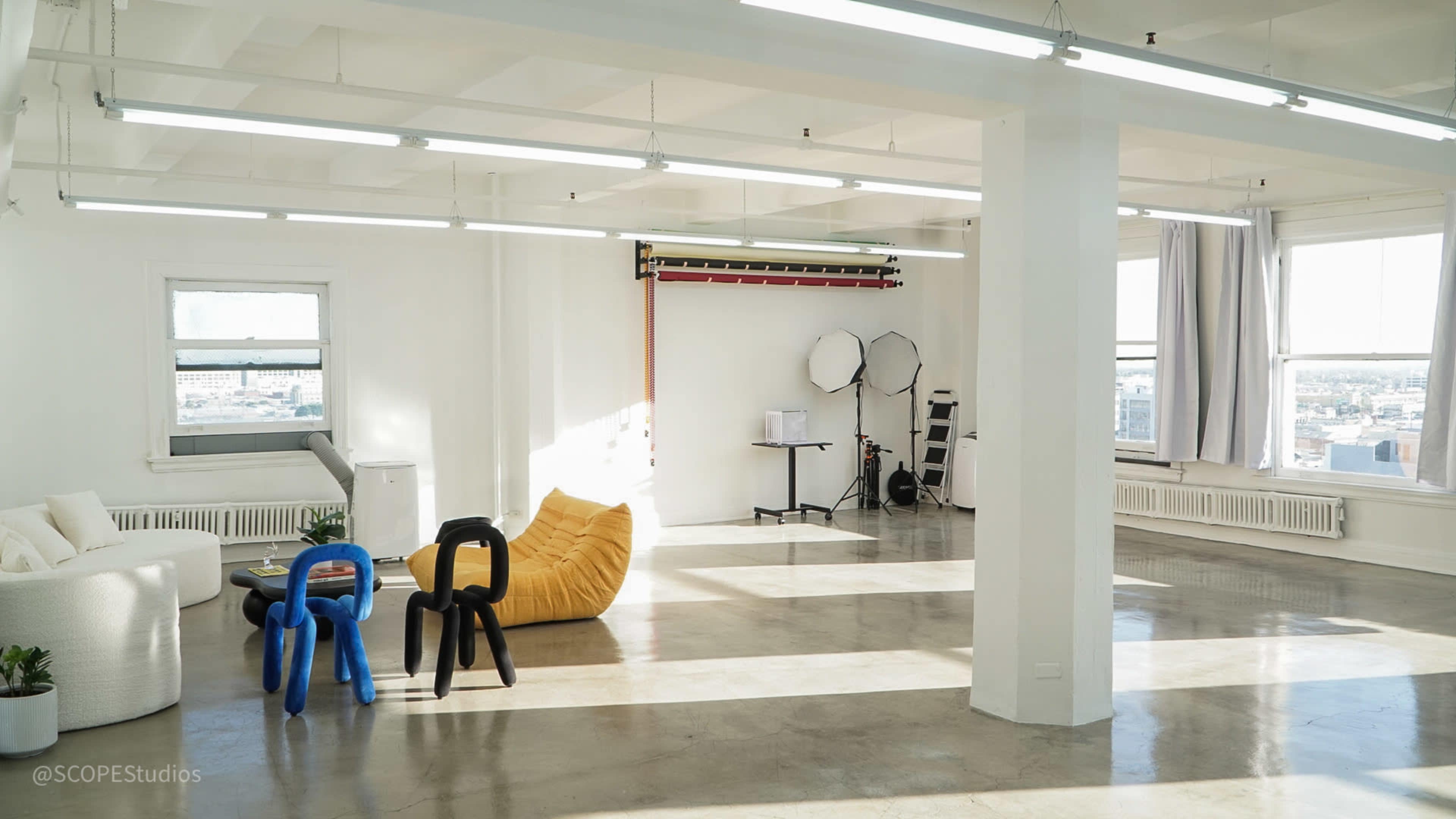 The image shows a spacious, well-lit studio with modern furniture, including a white couch, a yellow bean bag, and two colorful chairs, alongside photography equipment and large windows.