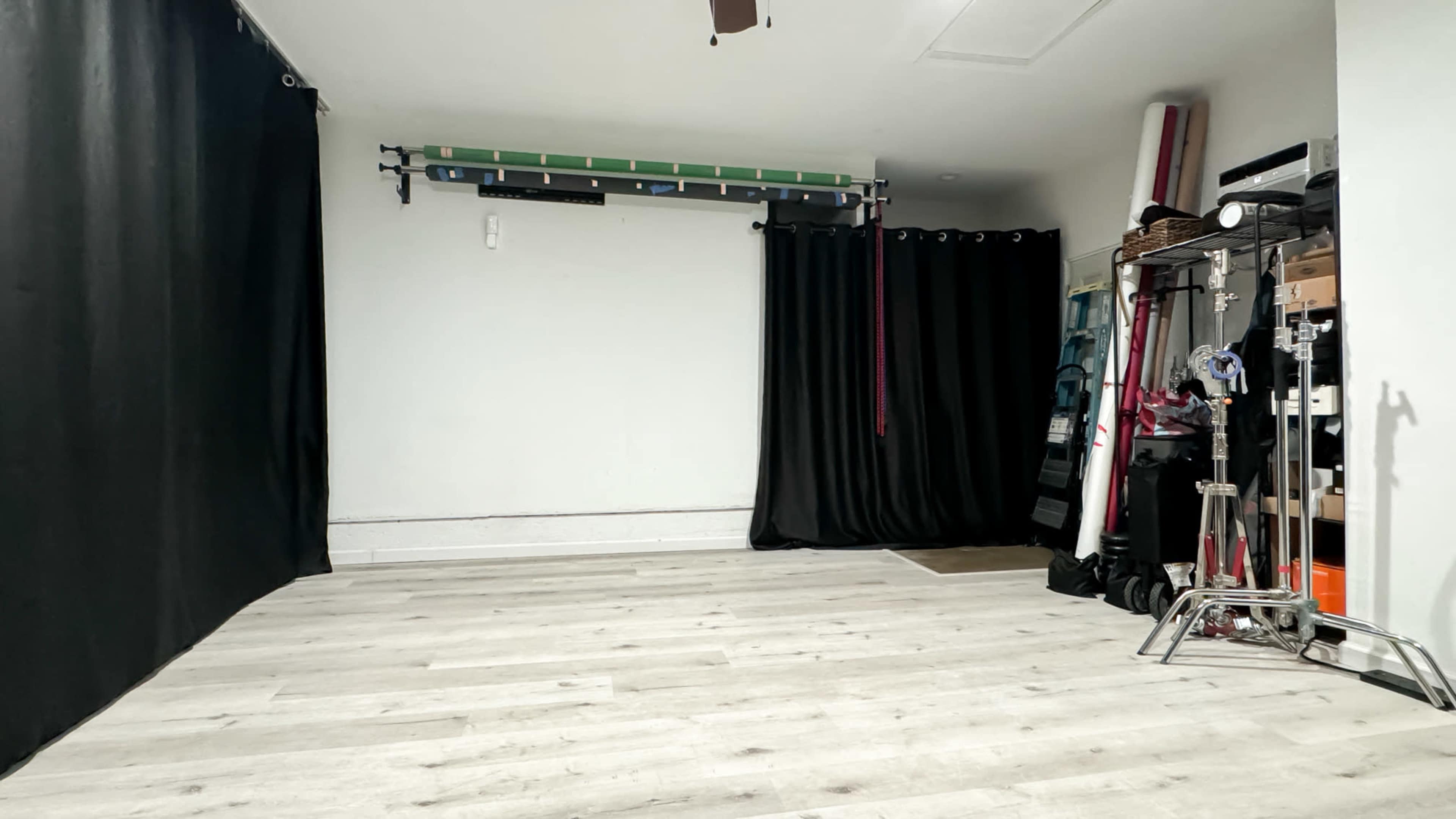 The image shows a minimalist indoor space with light wooden flooring, black curtains on one side, and various equipment and storage items against the wall.