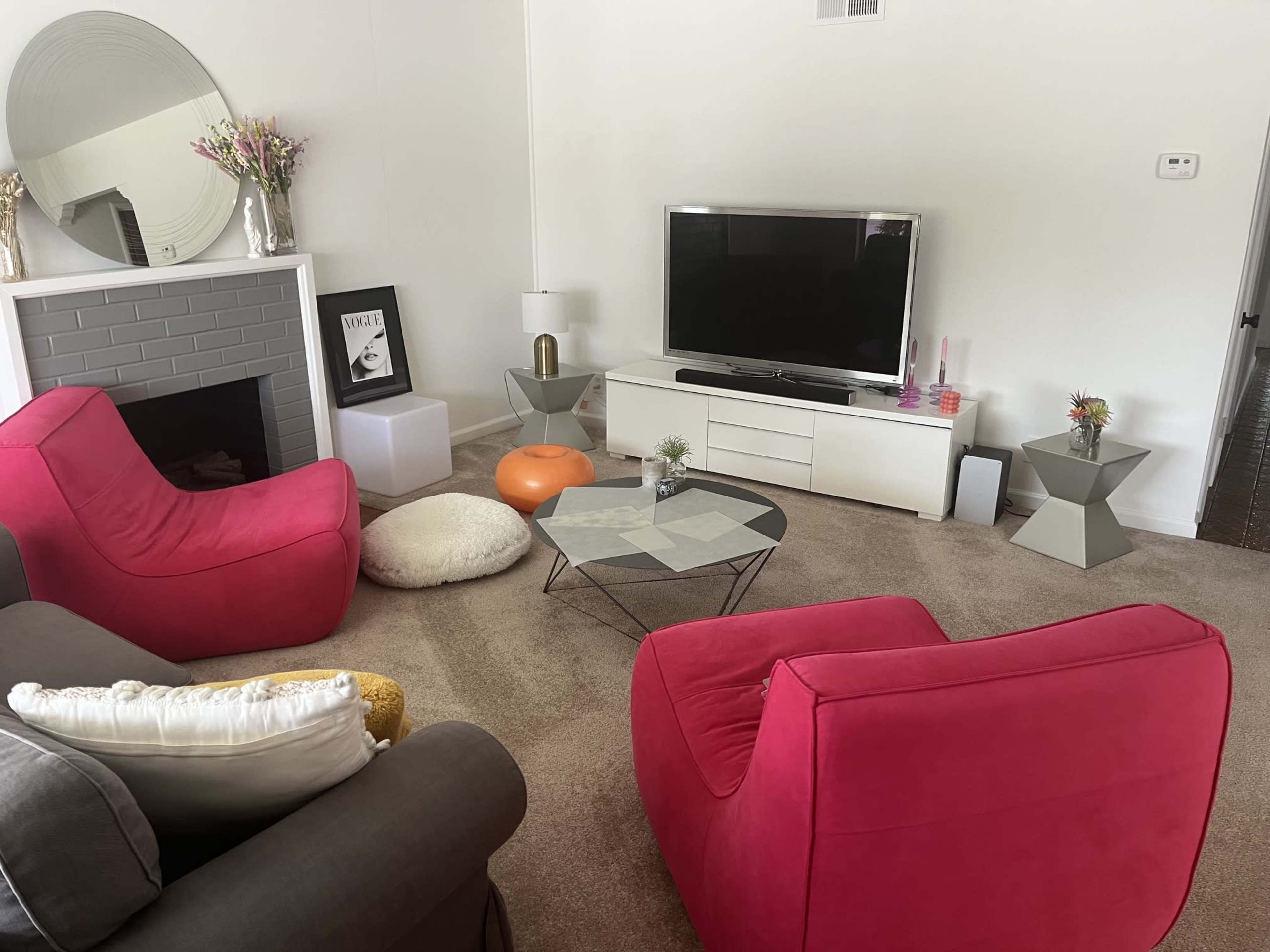 The living room features a modern setup with two pink chairs, a gray sofa, a coffee table, a television, and decorative elements like a round mirror and small vases.