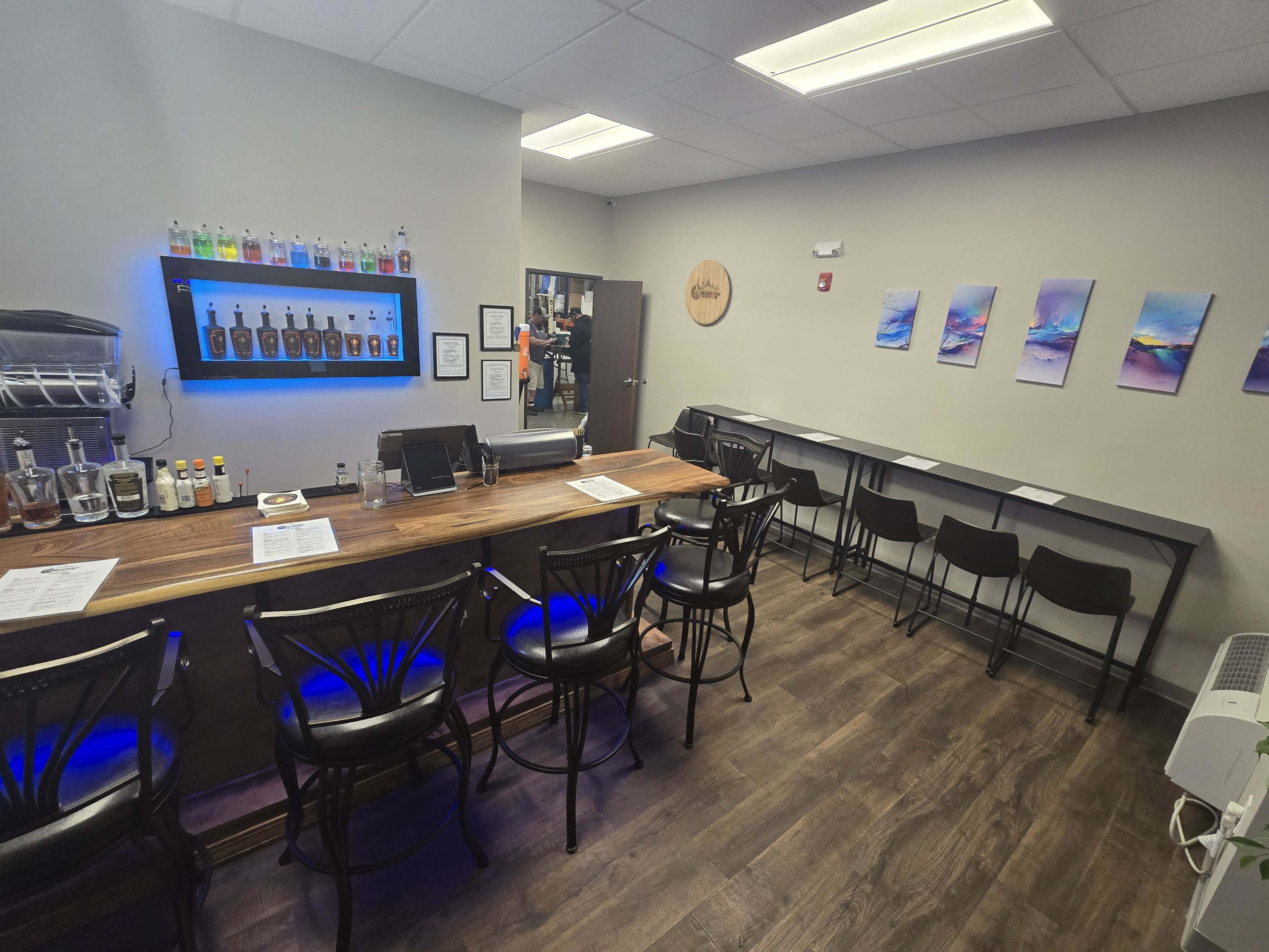 The image shows a small bar area with a countertop, a row of black chairs, and shelves displaying various bottles of liquor along one wall.